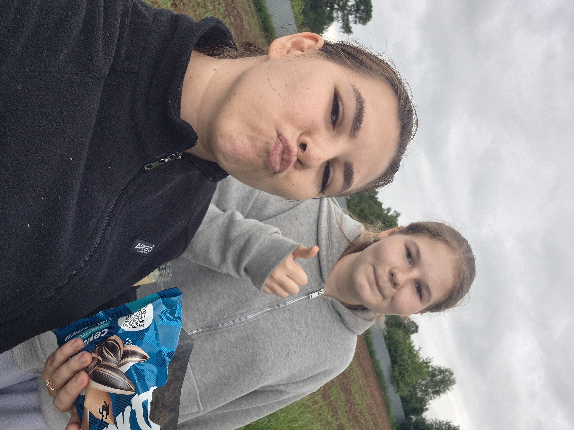 This image depicts two young women outdoors in Kharnavy, Russia, under an overcast sky. In the foreground, a young woman with brown hair, tied back, wears a black fleece jacket with an "Argo Classic" logo and a nose stud. She is making a pouting expression with her lips puckered. To her right and slightly behind, another young woman with lighter hair wears a grey zip-up hoodie. She holds a blue bag of sunflower seeds, featuring Russian text, a QR code, and images of seeds, in her left hand, and displays a thumbs-up gesture with her right hand. A small glass bottle with a light-colored liquid is visible near her left hand. The background features a brown, possibly tilled, field, a corrugated metal fence, and various green trees under a cloudy sky.