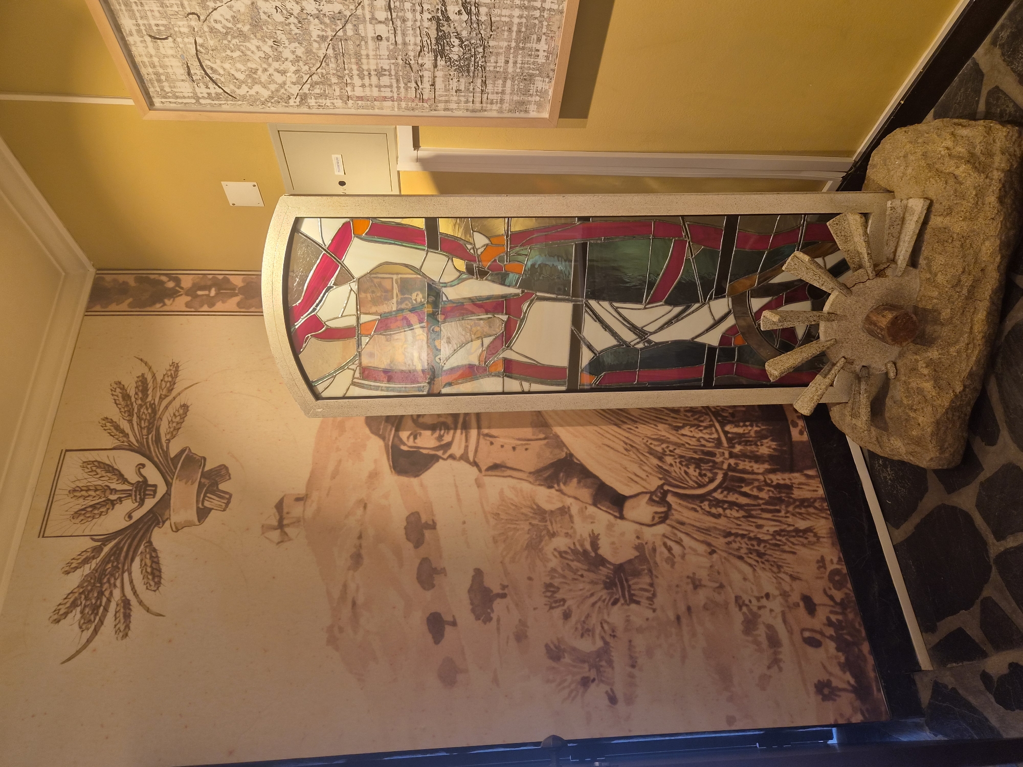 The image depicts an interior scene, likely within a museum or historical exhibition space, in Seia, Portugal. The dominant feature is a large mural on the left wall, rendered in a sepia tone. This mural illustrates a rural landscape with a windmill atop a hill in the background, and in the foreground, a woman in traditional attire, possibly a farmer or harvester, is depicted with a sickle amidst fields of wheat. Above the mural, a crest featuring wheat stalks and a shield is prominently displayed.
To the right of the mural stands a decorative stained-glass panel, tall and narrow, framed in a neutral-colored material. This panel features an abstract design with various colored glass pieces, including red, white, green, and orange, set against a lighter background.
In front of the stained-glass panel, resting on a large, rough-hewn stone, is a stone decorative element resembling a millstone or wheel with spokes. A small wooden piece is centrally embedded in this stone structure.
The floor is tiled with irregularly shaped dark grey stones. The walls are painted a warm yellow, with a white molding running along the top. To the right of the stained-glass panel, a light-colored electrical panel or utility box is mounted on the yellow wall, with a small white square plate above it. Further to the right, a framed artwork with a complex, abstract pattern, possibly a map or textile, is hung on the yellow wall. The overall impression is one of a culturally themed exhibit, possibly related to agriculture or local heritage in Seia.