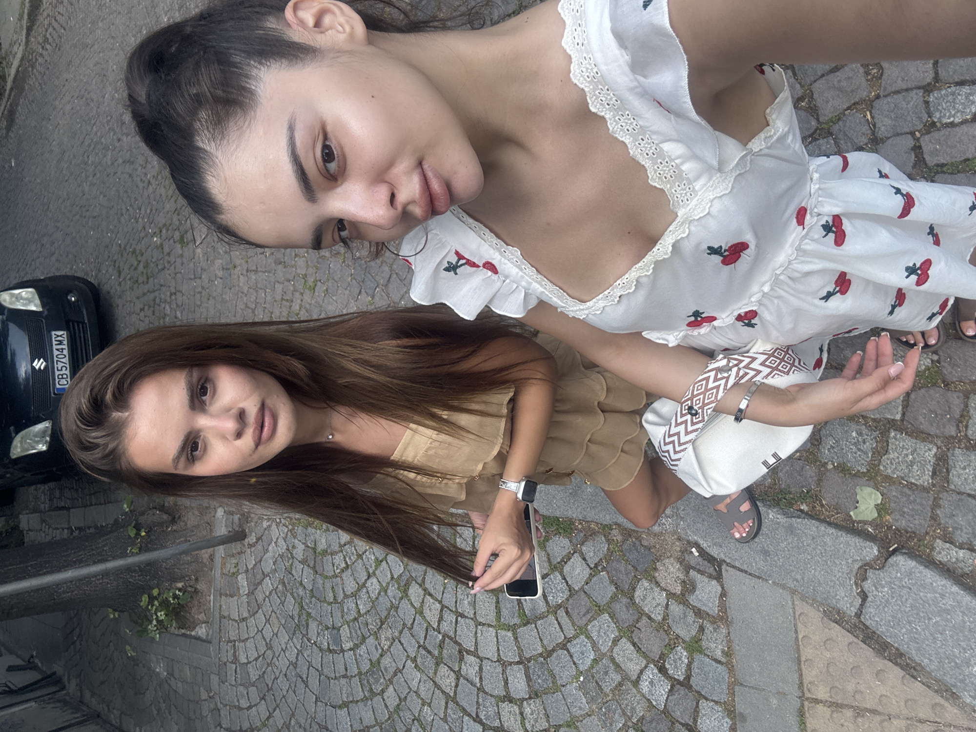 The image depicts two young women posing casually outdoors on a cobblestone street, likely in Plovdiv, Bulgaria, as specified. The photograph is taken from a slightly elevated angle and is presented rotated, showing the subjects and scene on their sides.
The woman positioned closer to the camera, seemingly taking the photo, has dark hair, possibly tied back, and is wearing a white dress. The dress features red cherry embroidery and ruffled lace trim along the neckline. She has a serene expression with a slight smile and is holding a white woven bag with a brown and white geometric pattern, on which a small "E" and a red heart are visible. She wears dark sandals.
Behind her, the second woman has long, straight brown hair and a neutral expression. She is dressed in a light beige or tan strapless top or dress with ruffled details. She wears a silver-banded watch on her left wrist and holds a white smartphone in her hand.
The setting is an urban environment characterized by a textured cobblestone street and irregular stone slabs. A black Suzuki car, displaying the Bulgarian license plate "CB 5704 MX," is parked in the background to the left. Small patches of green vegetation are visible growing between the stones, and a dark metal rail runs across the mid-ground. The natural, soft lighting indicates it is daytime. The overall atmosphere is relaxed and casual.