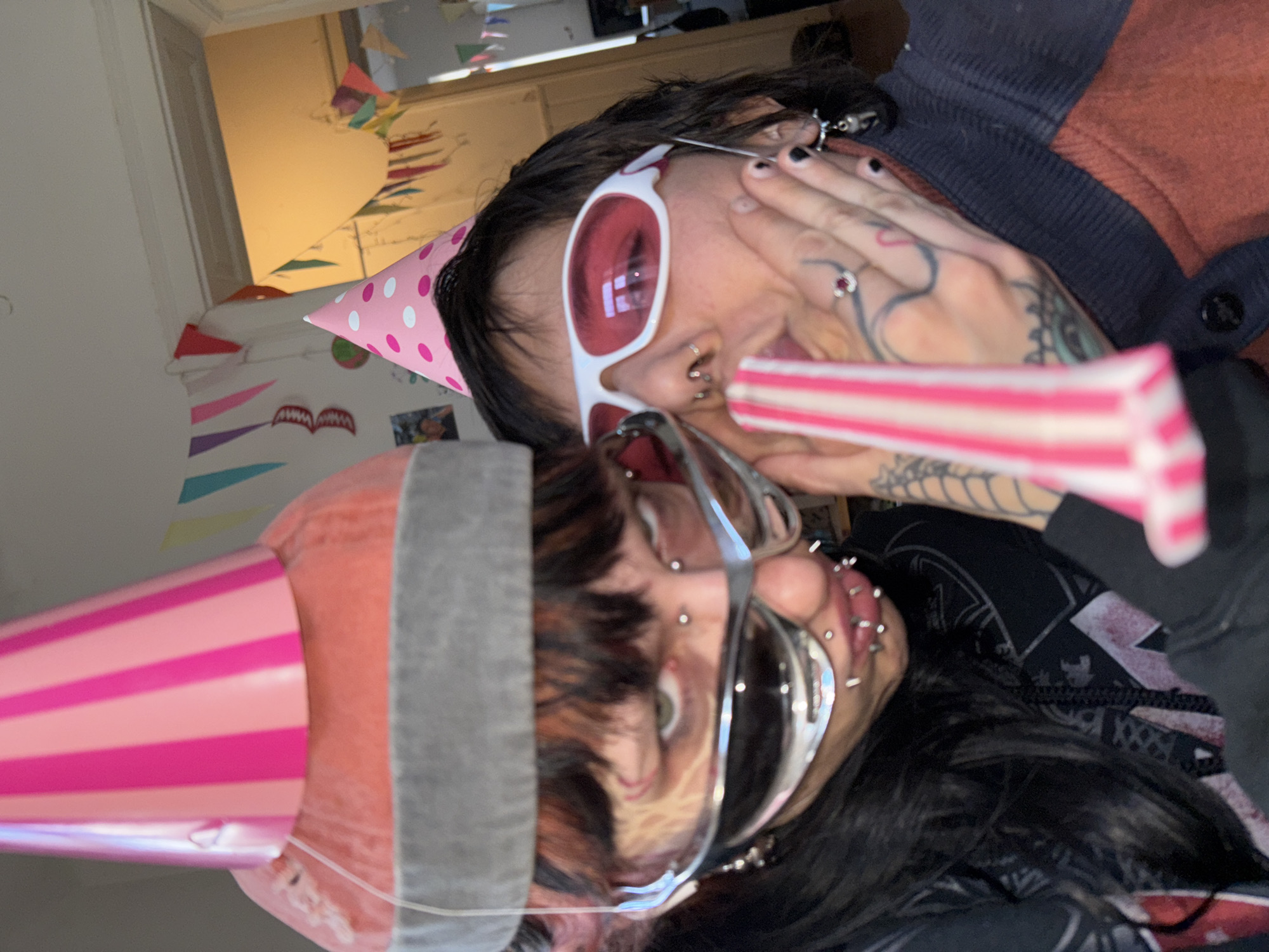 Two people are posing for a photo, likely indoors. One is wearing a pink party hat with white polka dots, white-framed sunglasses with red lenses, and is holding a pink and white party horn. The other person is wearing a pink and white striped party hat and silver-framed glasses. Both have piercings on their faces. The background includes a doorway, wall decorations, and what appears to be a room with streamers. The location is not identifiable as Sofia, Bulgaria from this image.