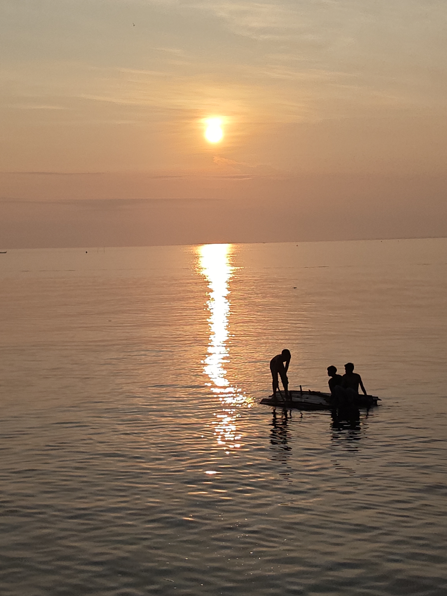 Three people are on a raft on the water. The sun is setting or rising. The city of Lhokseumawe, Indonesia, is not discernible in this image.