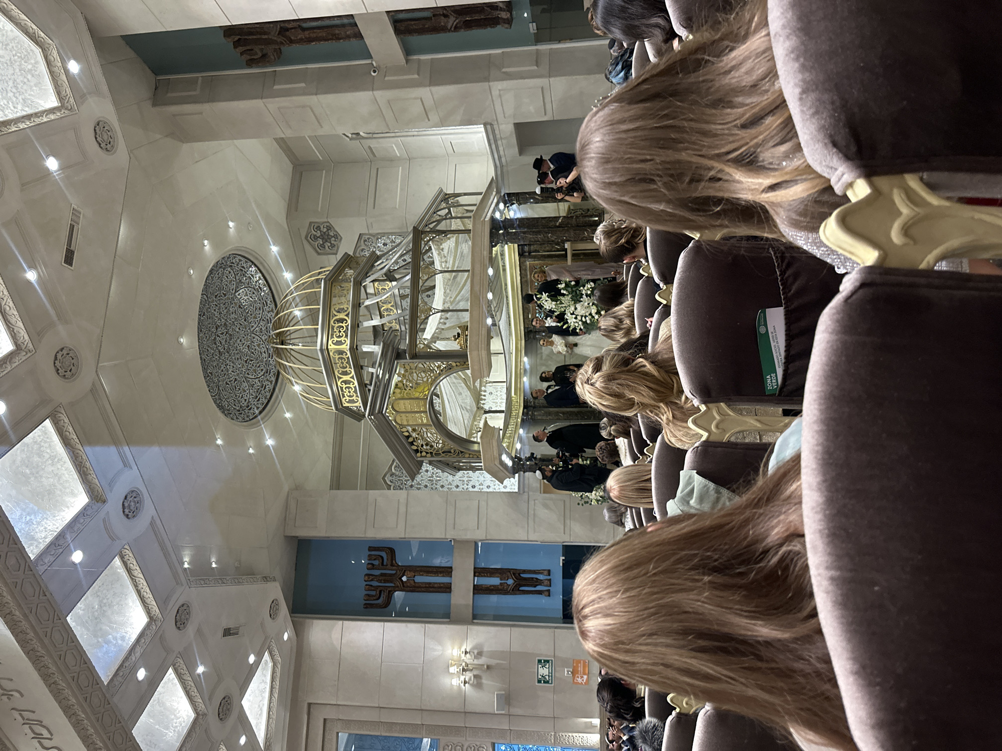 The image shows an interior view of a synagogue located in Mexico City, Mexico, as suggested by the program in the seat pocket. The perspective is from the rear of the seating area, facing the Bimah (raised platform). In the foreground, multiple rows of seats are visible, most of which are occupied by people, largely with the backs of their heads towards the camera. The Bimah is ornate and gold-colored, with a multi-tiered structure. A wedding ceremony appears to be in progress, with a white draped Chuppah visible atop the Bimah and figures dressed in dark suits and white dresses observed near the structure. Decorative elements adorn the walls, including a patterned circular design on the ceiling. A cross-like bronze design is on the wall behind. The lighting appears to be a mix of natural light from windows and artificial lighting from fixtures. The atmosphere is formal.