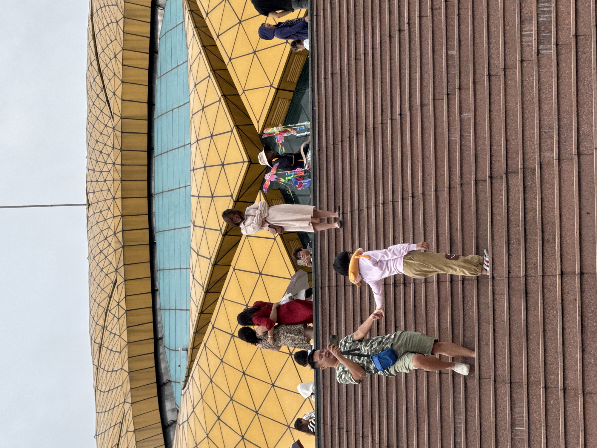 In Da Lat, Vietnam, a wide shot captures a bustling scene on a large, tiered staircase leading up to a distinctive yellow, geometric building with a blue-tinted glass section. On the steps, a father, wearing a camouflage t-shirt, khaki shorts, and a blue fanny pack, holds the hand of a child in a light pink long-sleeved shirt and khaki pants. The child has an orange neck pillow around their neck. The father is holding a smartphone, appearing to take a photo. Further up the stairs, a group of people are gathered. Two women, one in a floral dress and another in a red dress, are embracing. To the right, a woman in a light pink dress stands with her arms crossed. Another woman in a hat is selling colorful toys. Children and adults are visible throughout the scene, some looking at their phones, others in conversation. The weather appears overcast.