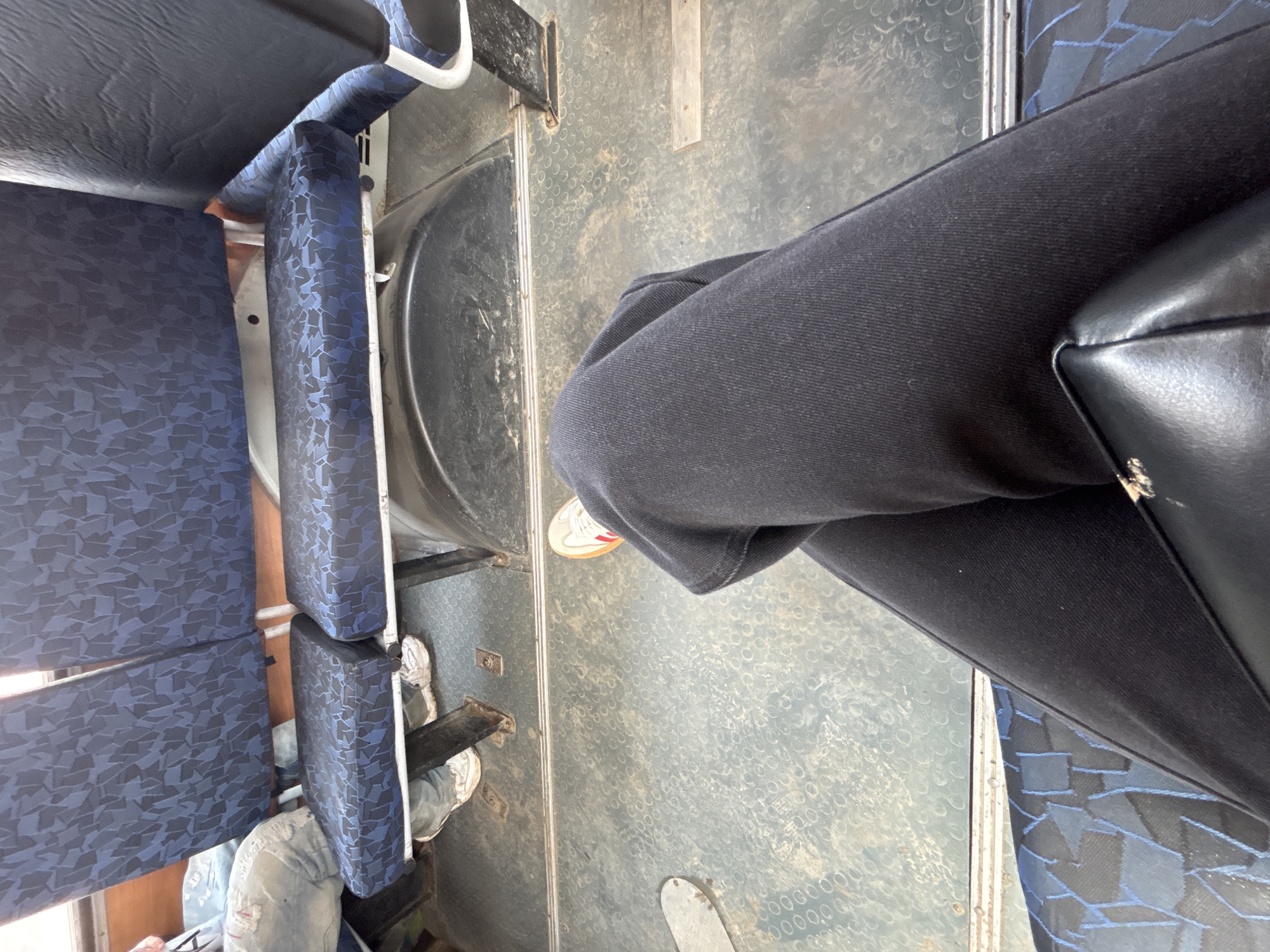The image is taken from an elevated perspective inside a bus or similar public transport vehicle. The frame is primarily occupied by seats with blue patterned upholstery, arranged in rows. A person, clad in black clothing, is seated, with their crossed legs and part of a black bag visible. The bus floor is grey and patterned. The location Фоки, Russia is unknown to be verifiable from the image.