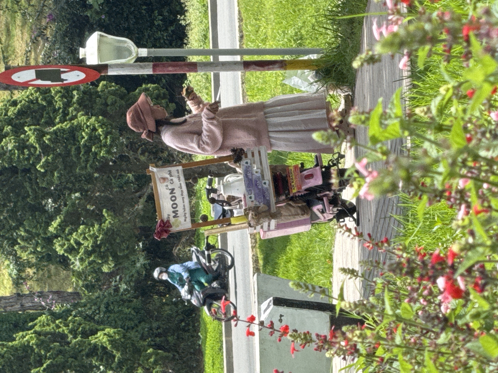 In Phường Xuân Hương - Đà Lạt, Vietnam, a woman in a pink sweater and a long white skirt stands on a wooden walkway next to a small mobile food cart. The cart is attached to a pink scooter and displays various items, including a sign that reads "MOON Cà phê" and smaller signs with Vietnamese text. The woman is holding a piece of food in her hand and appears to be eating it. Behind her, a street light and a traffic sign are visible. Further in the background, a person wearing a blue medical-style uniform rides a motorcycle. The environment is green and lush, with trees and grass, suggesting a park or roadside setting. In the foreground, red and pink flowers are in bloom.