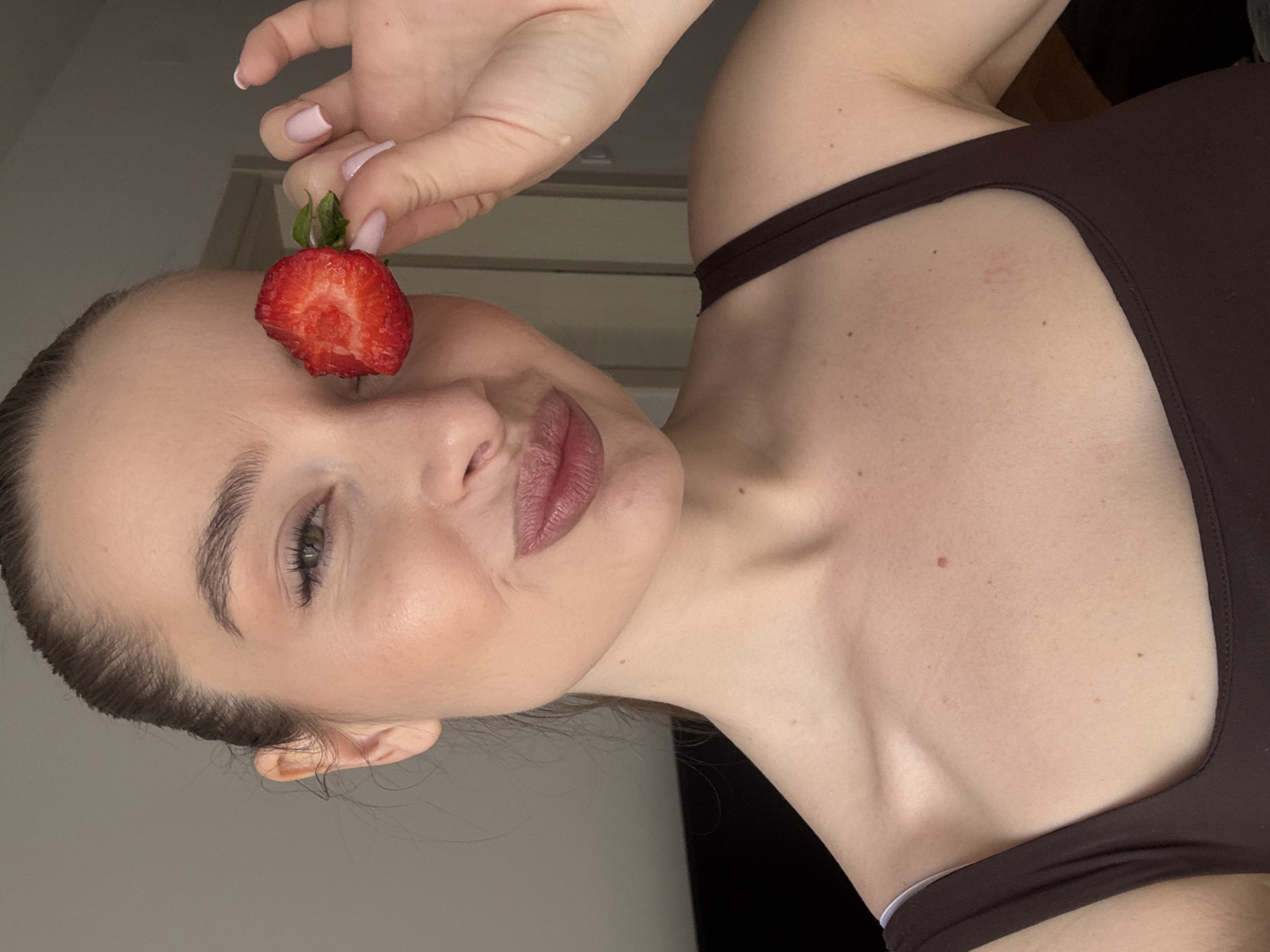 A portrait features a young woman captured from a tilted perspective, looking towards the viewer. She playfully holds a half-strawberry, obscuring her right eye with it.
The woman has light-toned skin, and her hair is neatly pulled back. Her visible left eye is a light color, framed by well-defined eyebrows and subtle eye makeup. Her lips are softly colored and slightly parted, forming a gentle pout or faint smile. Her right hand, with light pink nail polish, holds a vibrant red strawberry, sliced lengthwise, directly in front of her right eye. The prominent cut side of the strawberry displays its juicy, textured flesh.
She is wearing a dark brown top, likely a tank top or a one-shoulder design, which reveals her neck, collarbone, and a portion of her upper chest and shoulder. Her skin appears clear, with a few small moles naturally present. The background is a simple, light-colored wall, indicating an indoor setting. The soft, even lighting suggests it is daytime. The overall expression and composition convey a lighthearted and gentle mood. No visible text is present in the image. This scene takes place in Budva, Montenegro.