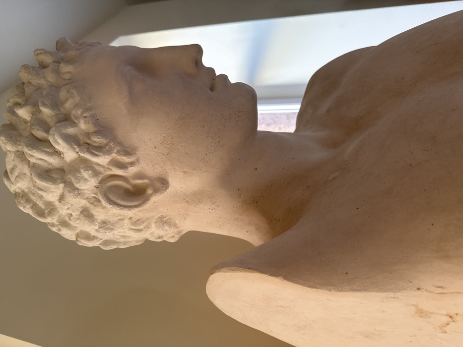 A light-colored, possibly plaster or cast stone, male bust is depicted in a three-quarter profile, facing right, within an indoor setting in Talin, Armenia. The upper torso, neck, and head of the figure are visible. Features include short, curly hair, a distinctly sculpted right ear, a prominent straight nose, and undetailed eye sockets. The mouth is slightly open. The material is off-white to cream with a matte, textured finish and scattered dark flecks. A sculpted element, appearing wing-like or as drapery, originates from the figure's upper left back/shoulder area, extending upwards. The background on the left consists of a plain, light-toned wall. The background on the right reveals a vertical window providing ambient light. No individuals or actions are present.