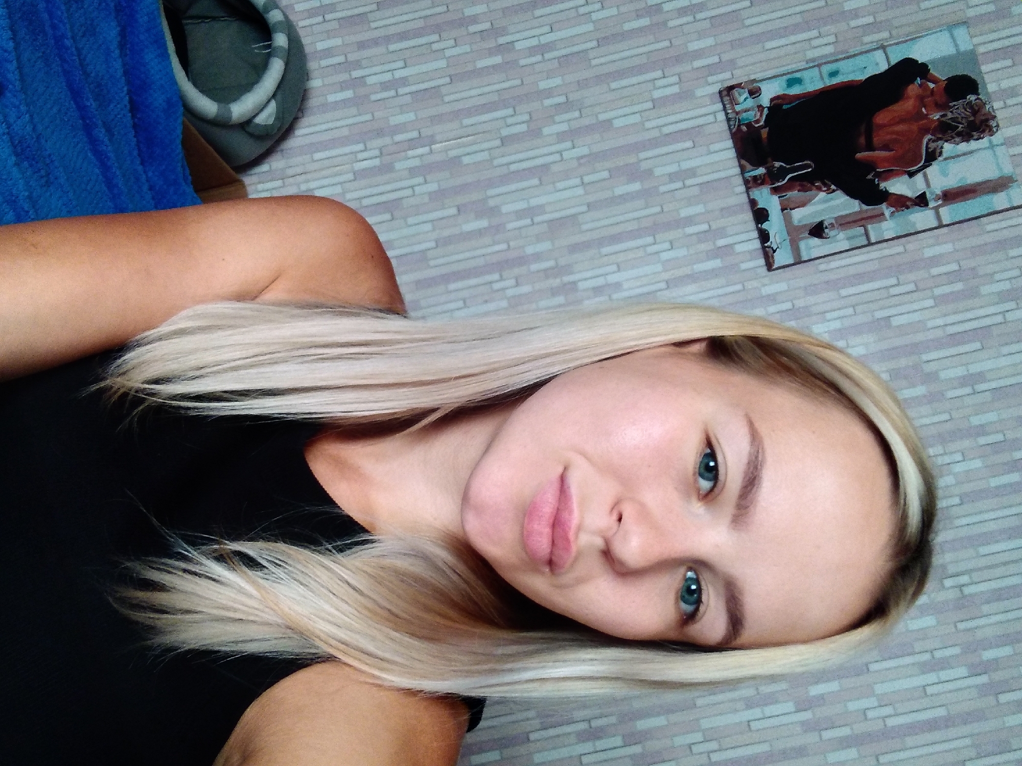 A young woman with blonde hair and blue eyes is looking at the camera. She is wearing a black top. She is indoors, in front of a wall with a patterned wallpaper. To the left of the woman, there is a framed picture hanging on the wall, depicting a couple holding wine glasses. In the lower left corner of the image, a blue blanket and a part of a pet bed are visible. The exact location in the city of Radisne, Ukraine cannot be determined from the image.