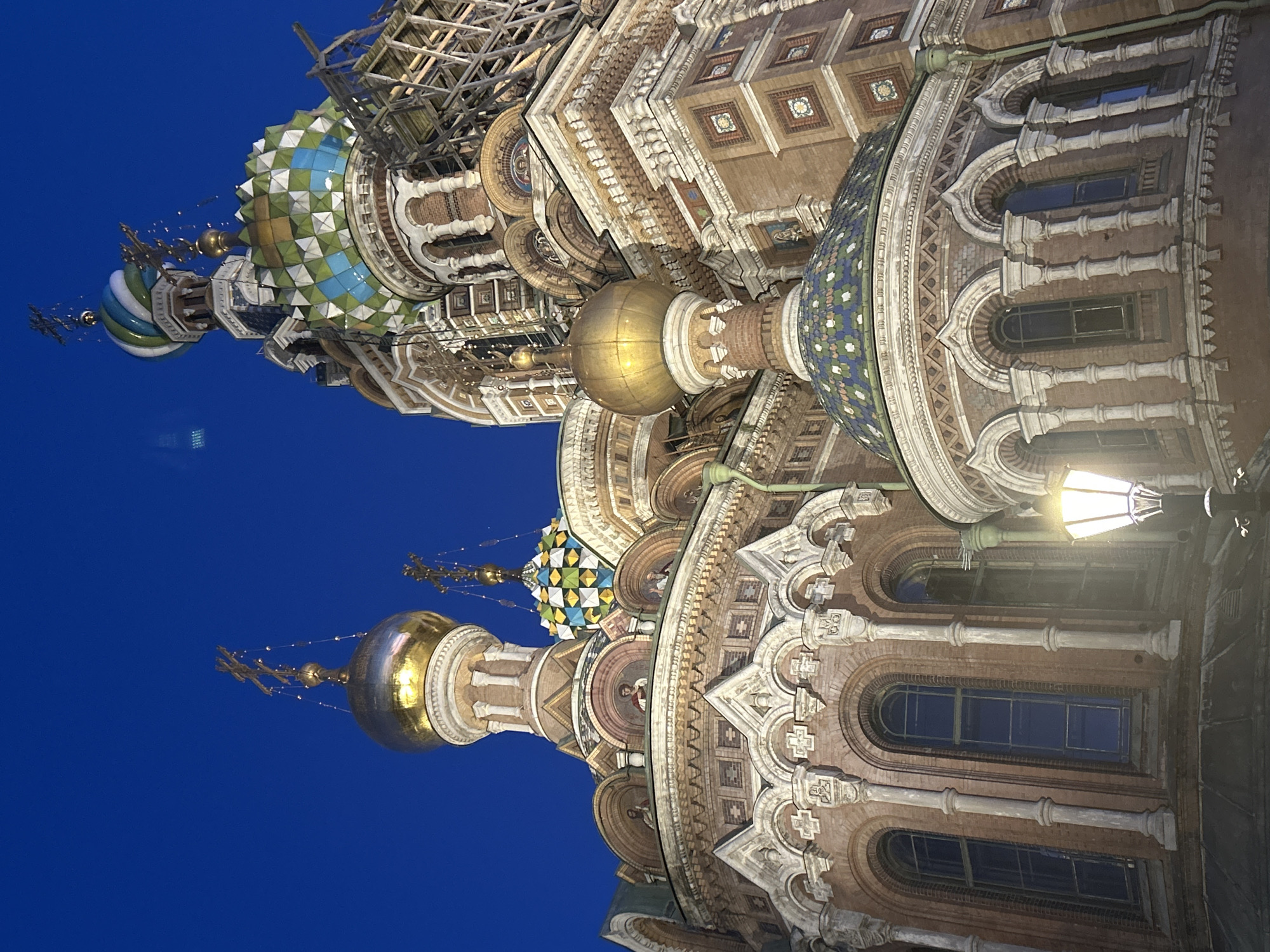 The image captures a low-angle view of the Church of the Savior on Spilled Blood in Saint Petersburg, Russia, at dusk. The sky is a deep, clear blue, indicating the transition from day to night. The iconic onion domes of the church are the primary subjects, with one prominent golden dome catching the artificial light and reflecting it. Another dome, adorned with colorful mosaic tiles in green, blue, and white diamond patterns, is visible, along with scaffolding on one side, suggesting ongoing restoration or construction work.
The intricate facade of the church is visible, characterized by its red brickwork, white decorative elements, and arched windows. Small mosaic portraits are embedded within some of the upper architectural features. A vintage-style street lamp with a glowing bulb is positioned in the lower right of the frame, illuminating the foreground and adding to the atmospheric lighting of the scene. The overall impression is one of grandeur and historical significance, highlighted by the architectural details and the twilight setting. No people are visible in the image.