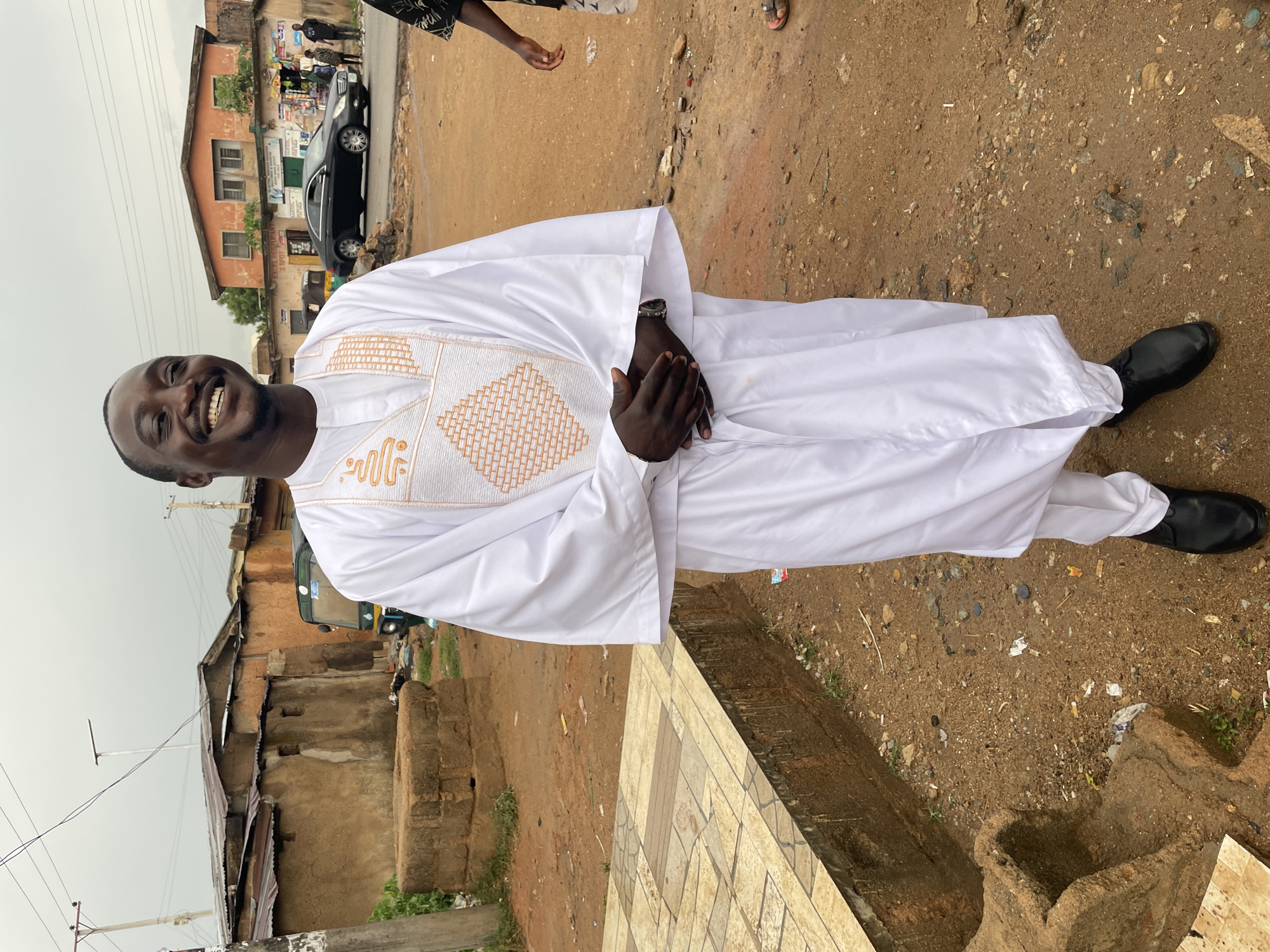 A medium shot captures a smiling man in a long, white, embroidered garment, positioned in what appears to be an urban area of Jos, Nigeria. The surroundings suggest a relatively underdeveloped part of the city, with a mix of buildings ranging from a two-story structure with an orange facade to more rudimentary, single-story dwellings with corrugated iron roofs and weathered walls. The ground is uneven and consists of dirt, gravel, and scattered debris. The subject is standing near a tiled surface, possibly a curb or sidewalk. A black car is visible in the background, along with indistinct figures near a shop or roadside stall. The sky is overcast, indicating soft, diffused lighting.