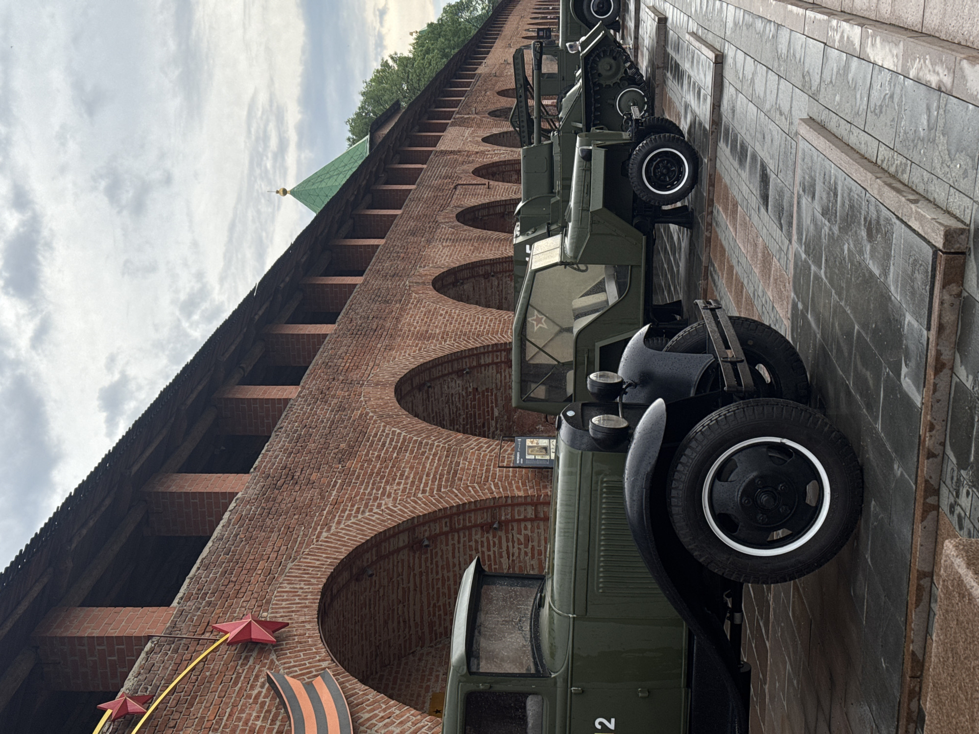 This image shows a view of the Kremlin wall in Nizhny Novgorod, Russia, with several vintage military vehicles parked along its base. The brick wall features arched openings and red star decorations. The sky is overcast.