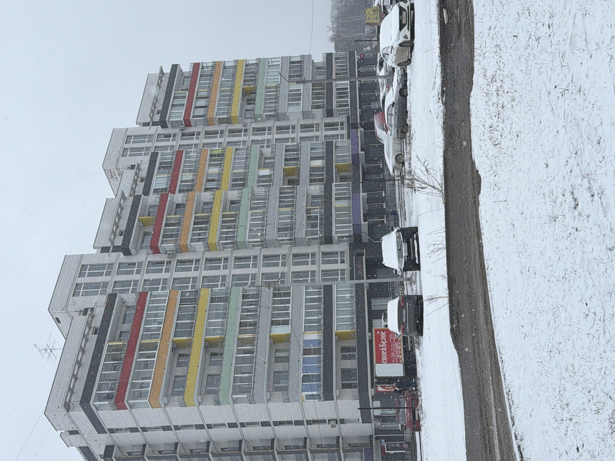 The image depicts a snowy street scene in what appears to be the city of Chaikovsky, Russia. A multi-story residential building with a brick facade is visible. The building has sections of red, yellow, green, and blue accent colors. Cars are parked along the street, some covered in snow. The road and sidewalk are covered in a layer of snow. The sky is overcast, and snow is falling. A truck is parked on the street.
