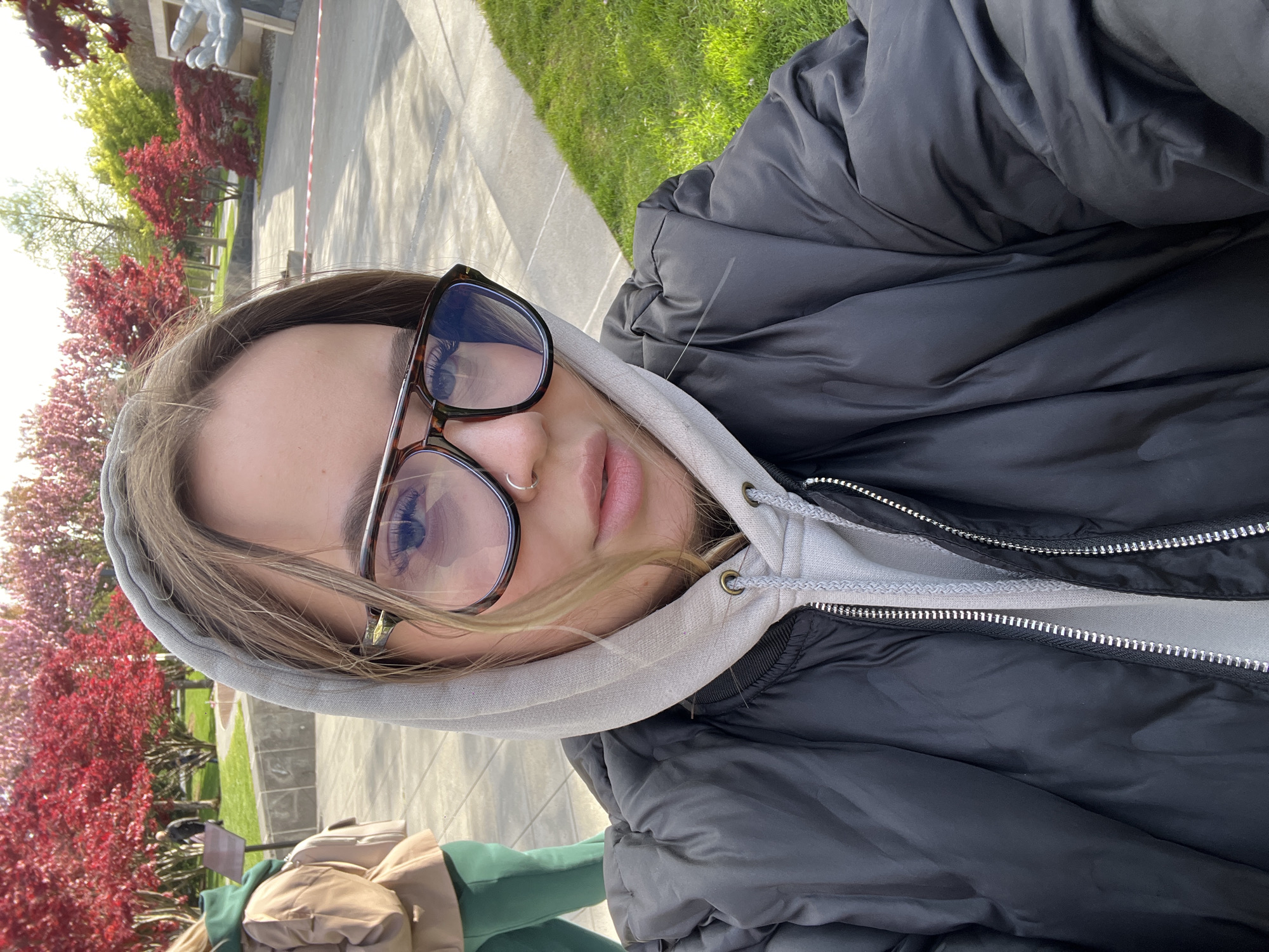 A young woman is featured in a close-up selfie taken outdoors in what appears to be a park in Krasnodar, Russia. She is positioned centrally, looking towards the camera with a neutral expression. Her light brown hair is visible beneath a light grey hooded sweatshirt, which is worn under a black puffer jacket. She is wearing large, dark-rimmed glasses and a small silver nose ring.
The background reveals a bright outdoor environment. To the left and behind her, numerous trees with vibrant red or pinkish foliage suggest either spring blossoms or autumn colors. A paved path stretches through the scene, bordered by green grass. In the far background, a large grey sculpture, possibly depicting a human hand, is partially visible, along with other distant greenery and structures. Another individual, partially visible in the lower left, is wearing a green top and has a beige bag resting near them.
The lighting is bright and clear, indicating a sunny day, likely during midday or early afternoon, with distinct shadows visible on the ground. No visible text is present in the image. The overall scene captures a casual moment in a picturesque park setting.