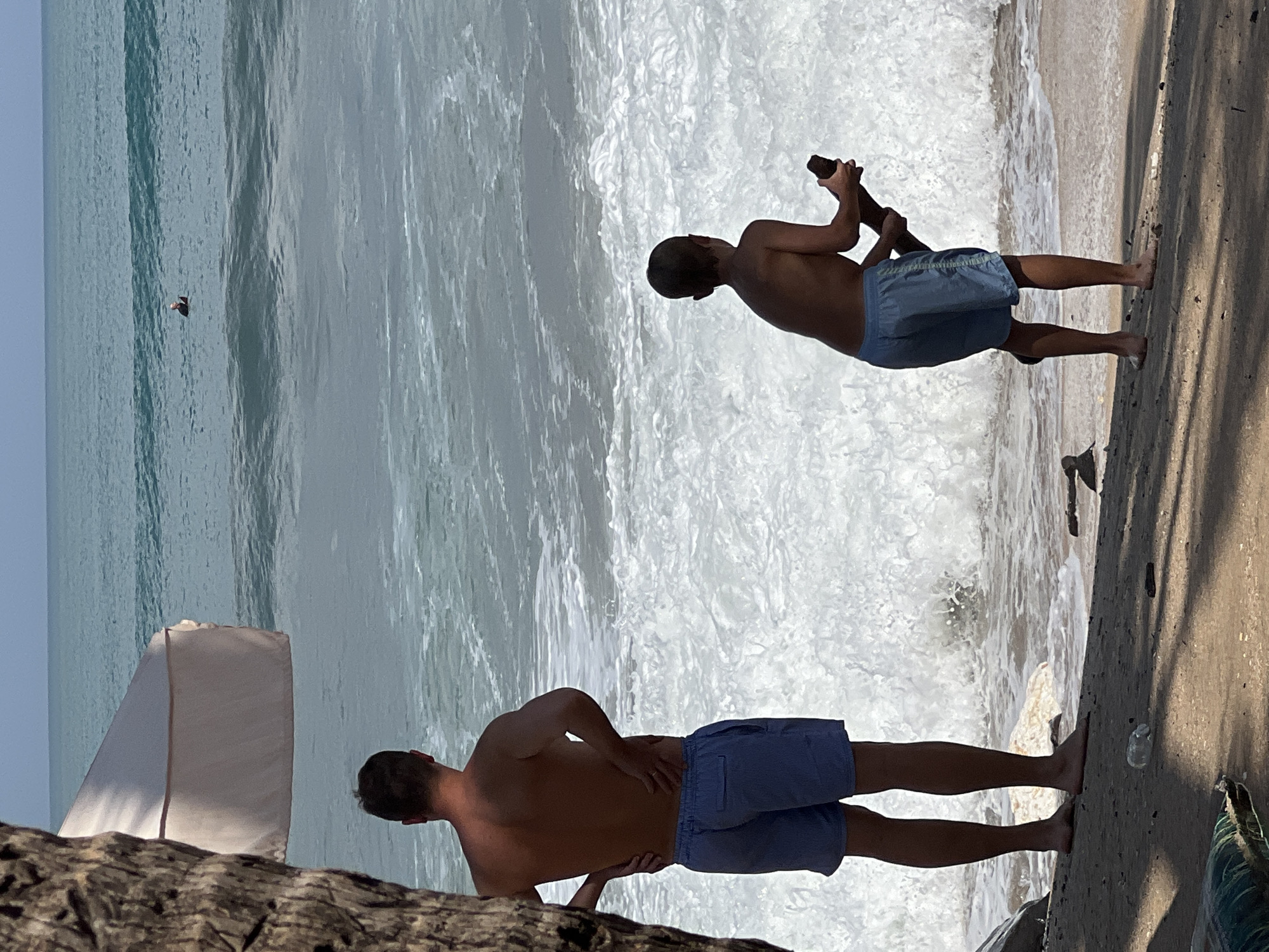 A medium shot captures a sandy beach in Kuta, Indonesia, where two individuals stand near the water's edge. On the left, a shirtless man wearing blue striped swim shorts stands with his back to the camera, hands on his hips. To the right, a young boy, also shirtless and wearing blue swim shorts, holds a piece of driftwood. The ocean in front of them is active with crashing waves and white foam. In the background, a lone person can be seen in the water. A large umbrella shades part of the beach in the upper left quadrant, and a palm tree trunk is visible on the far left. The sandy shore is scattered with small debris, including a plastic bottle.