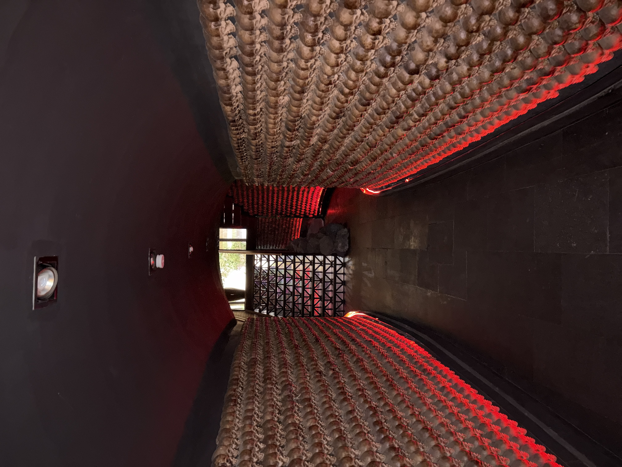 The image depicts a corridor or tunnel in what appears to be the Templo Mayor museum in Mexico City, Mexico. The walls are dark and slightly curved. On both sides of the corridor, arrays of skulls are visible, possibly part of a tzompantli display. These skulls are illuminated with red lighting. At the end of the corridor, daylight is visible through a window behind a patterned metal grate. A pile of rocks or artifacts is visible behind the grate. Speakers are mounted on the left wall. The floor is dark and paved.