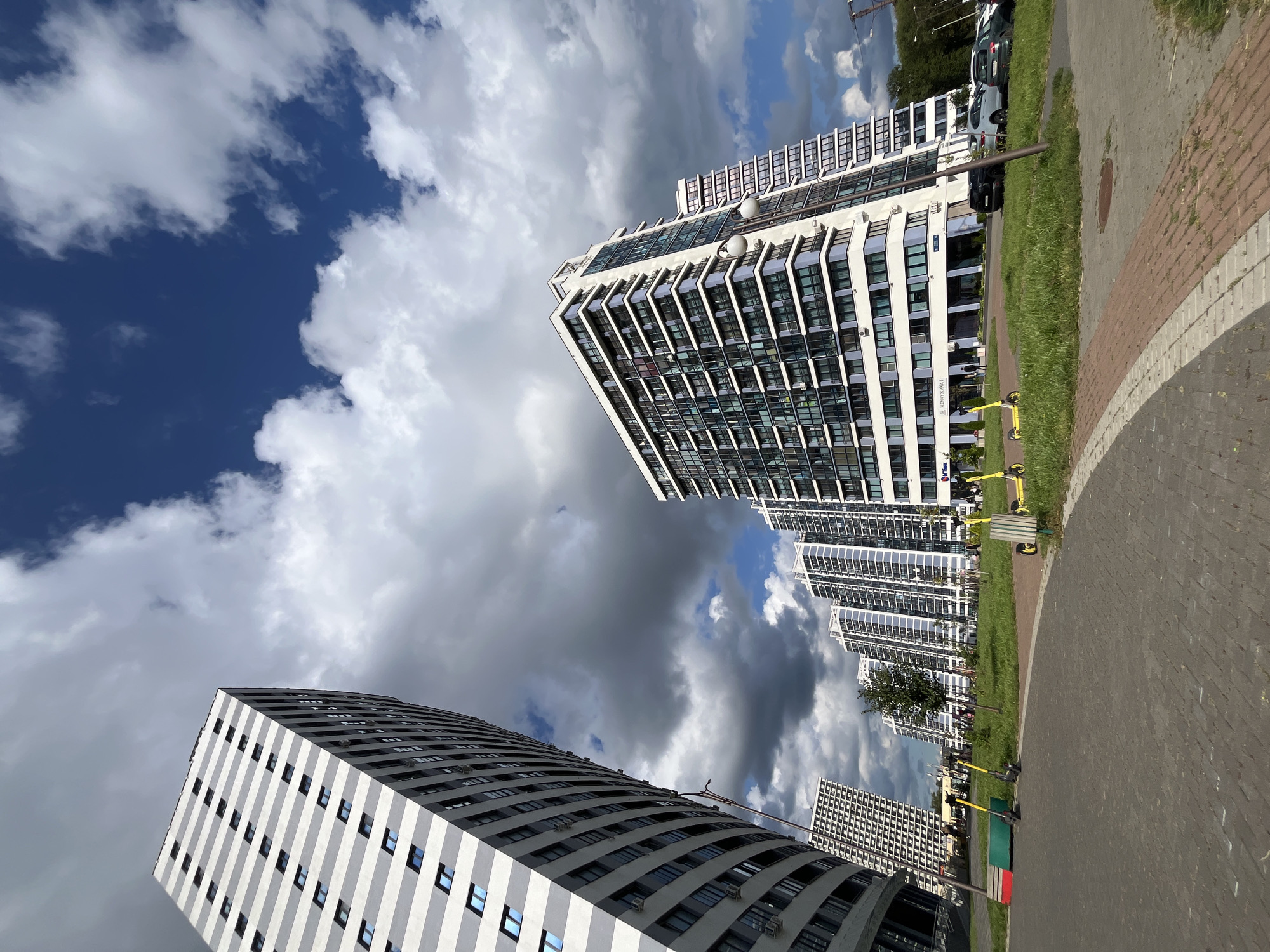 The image shows a street view of a modern residential complex in Minsk, Belarus, under a cloudy sky with patches of blue. The architecture is characterized by tall, contemporary buildings with white and grey facades and numerous windows. In the foreground, a paved area with brick accents leads to a grassy strip where several yellow electric scooters are parked. Beyond the green space, a series of apartment buildings are visible, varying in height and design. One building on the left has a distinctive curved facade and a striped pattern of grey and white. Another prominent building in the center is angular, with a facade featuring alternating white bands and rows of windows. Further back, more buildings of similar style stretch into the distance. Cars are parked along the side of the road in front of some of the buildings. A few individuals can be seen walking or standing near the entrance of one of the buildings. The overall impression is one of urban development with a focus on modern living spaces.