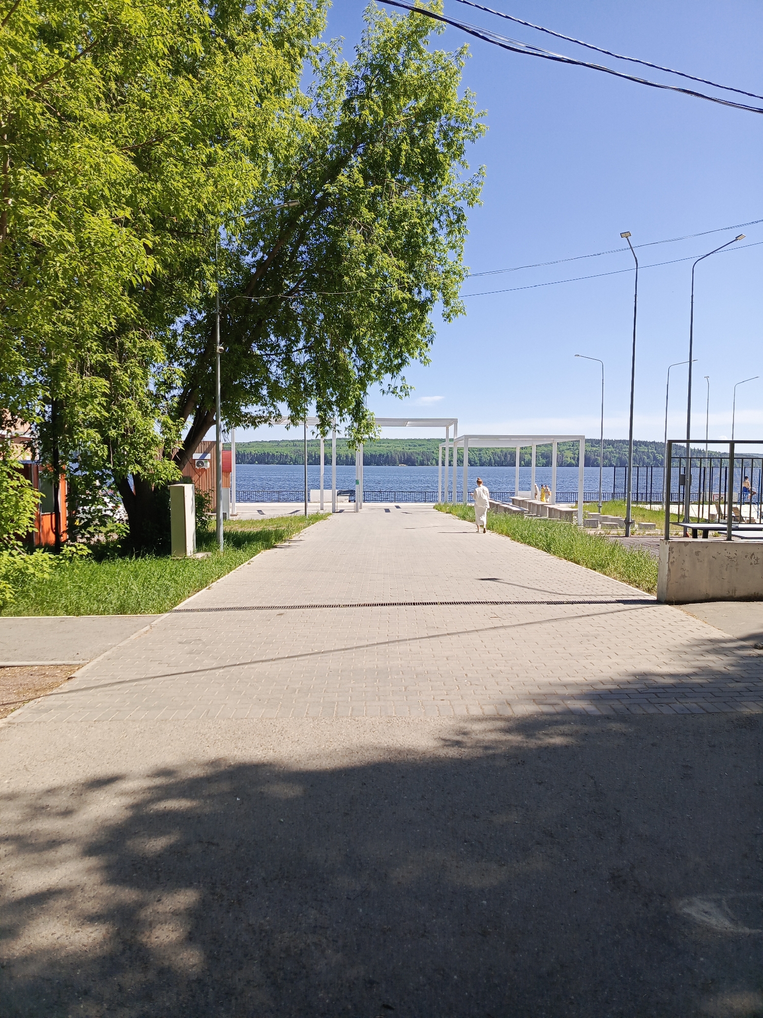 The image shows a paved walkway leading towards a waterfront area in the city of Chaikovsky, Russia. The path is flanked by greenery on the left and a concrete barrier and park benches on the right. In the distance, a white pergola structure is visible near the water's edge. A person is walking away from the camera on the path. A large tree with dense green foliage stands to the left of the walkway. Overhead, power lines stretch across the blue sky. The foreground is shadowed, likely by trees not visible in the frame.