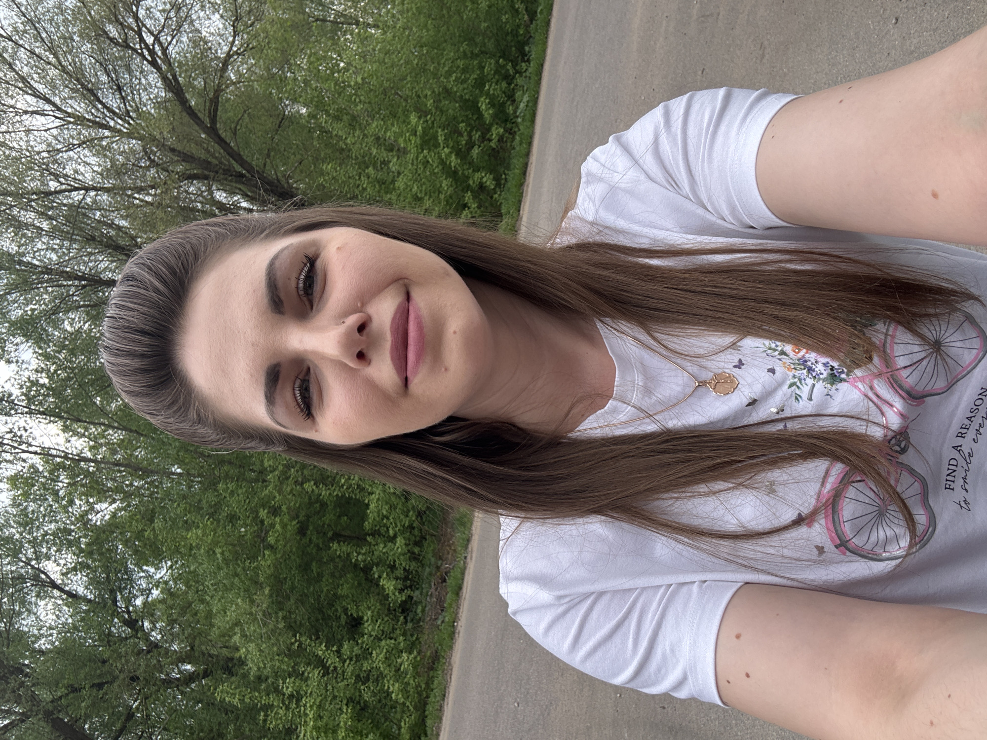 A young woman is taking a selfie outdoors. She has brown hair, fair skin, and is wearing a white T-shirt with a bicycle design on it. The background shows green trees and a paved path. It is daytime. There is no indication of the location being Shevlyakovo, Russia, in the image.