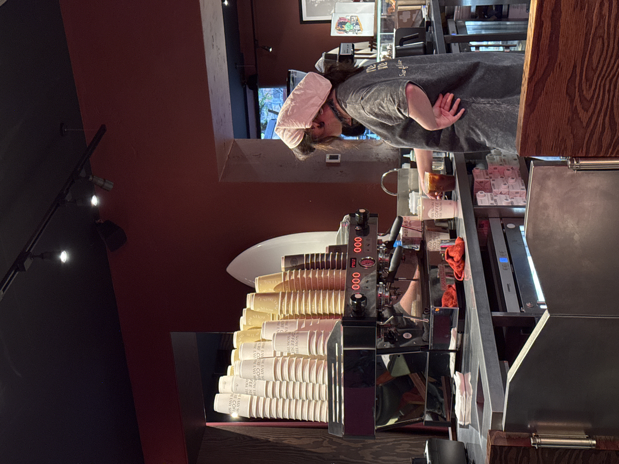 The image captures a scene inside a cafe or coffee shop, likely in Yerevan, Armenia, though without definitive elements to ascertain the location. A woman is working behind the counter, apparently in the act of preparing a drink. She has short brown hair, and is wearing a grey shirt with the text "Surflict" and a pink towel-like wrap on her head. Her attention is focused on the drink preparation in front of her. The counter is equipped with a professional espresso machine, stacked with cups of "MOI KOI EASY SLOW" branding. There are also small cartons, possibly milk or juice, arranged in neat rows. The environment has both light and dark areas; the red wall on one side contrasted with the dark ceiling and lighting fixtures. The cafe's general decor seems casual and slightly modern.