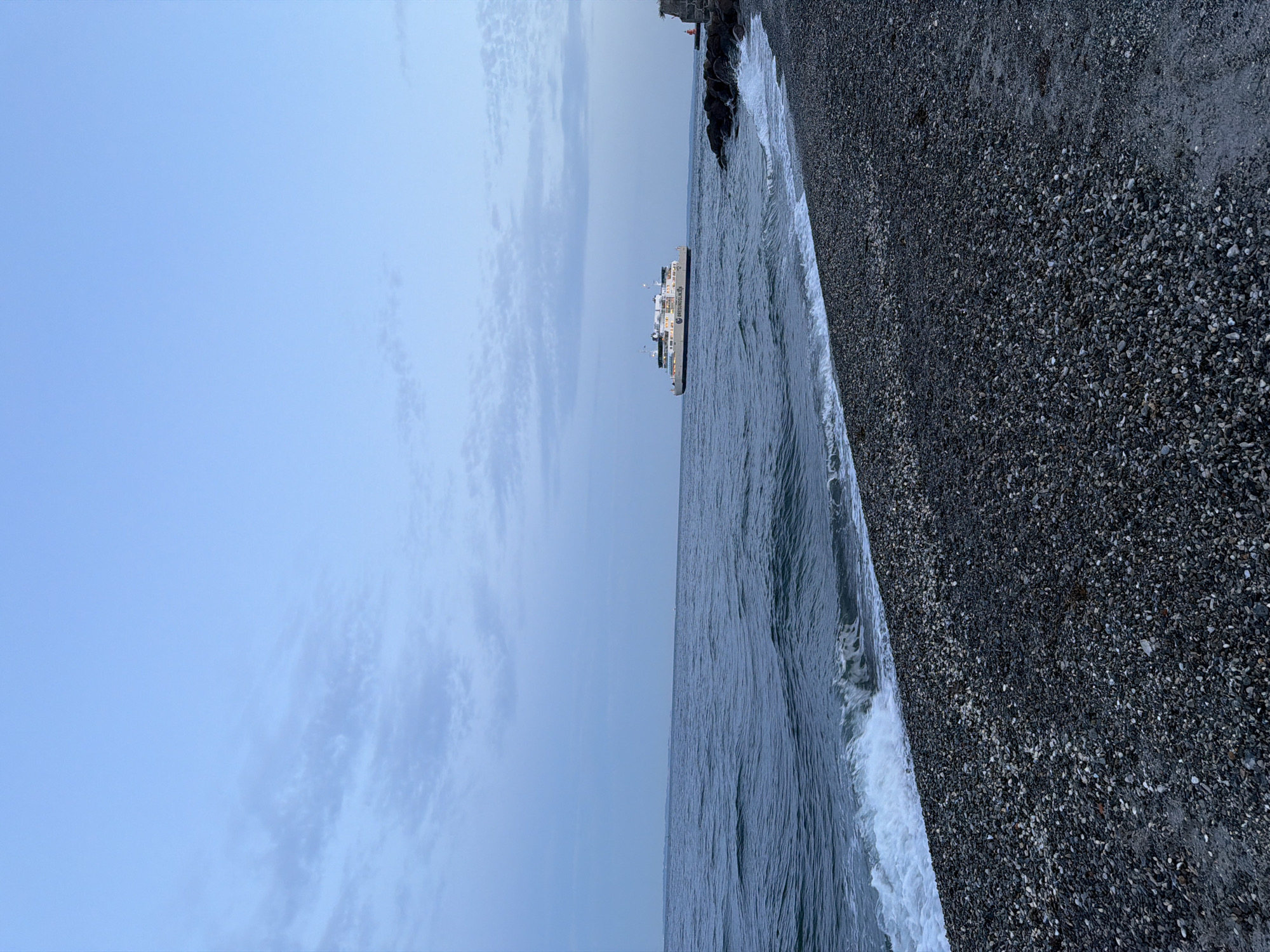 An image depicts a serene seascape with a ferry navigating the waters of Elsinore, Denmark. The sky is a pale blue with wispy clouds, suggesting either early morning or late evening. The sea is a deep blue, with gentle waves lapping against a pebble beach in the foreground. The beach is covered with an assortment of dark and light-colored pebbles. In the distance, a large, white ferry with the text "ØRESUNDSLINJEN" is visible, sailing horizontally across the frame. To the right of the ferry, a rocky structure juts out into the sea, with a small red beacon on top. The overall atmosphere is tranquil and evokes a sense of calm.
