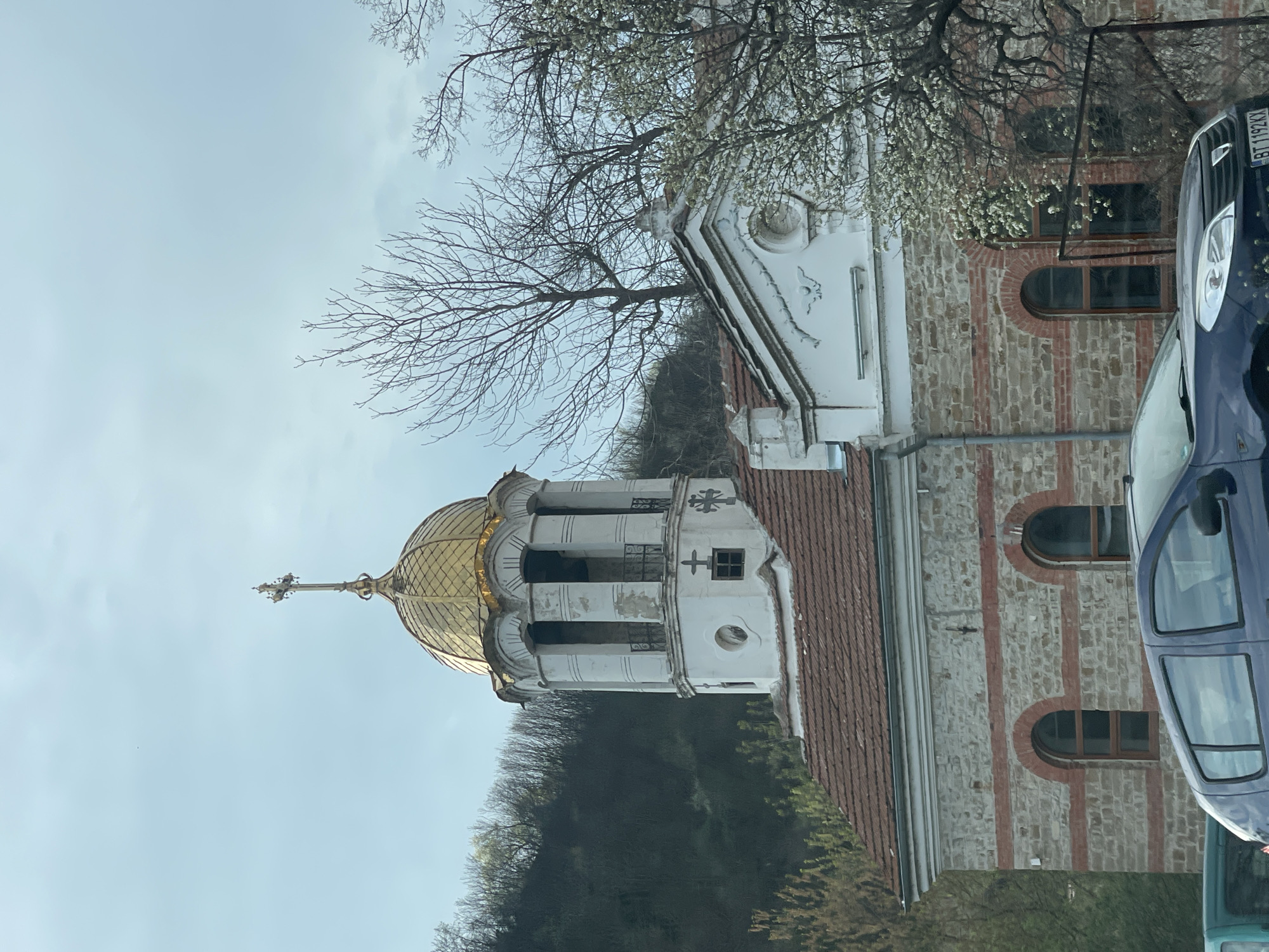 The image depicts the Sts. Cyril and Methodius Church in Veliko Tarnovo, Bulgaria. The building is constructed from stone and brick, with white architectural details. The church's golden dome is topped with a cross. A white bell tower stands next to the dome. A tree with bare branches partially obscures the upper portion of the building. A dark blue car is parked partially in front of the church. The sky is overcast. The building sits on a hill among trees.