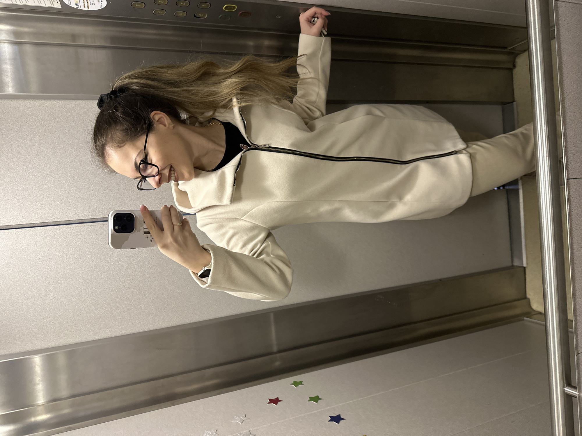 A woman is taking a selfie in an elevator. She has her hair in a ponytail with a black scrunchie. She is wearing glasses and a cream-colored tracksuit with a black top underneath. She is holding a white smartphone in her left hand. The elevator has a stainless steel interior, with control buttons on the upper panel. There are colorful star stickers on the floor of the elevator. The location is not explicitly stated in the image and therefore not known to be in the city of Stara Zagora, Bulgaria.