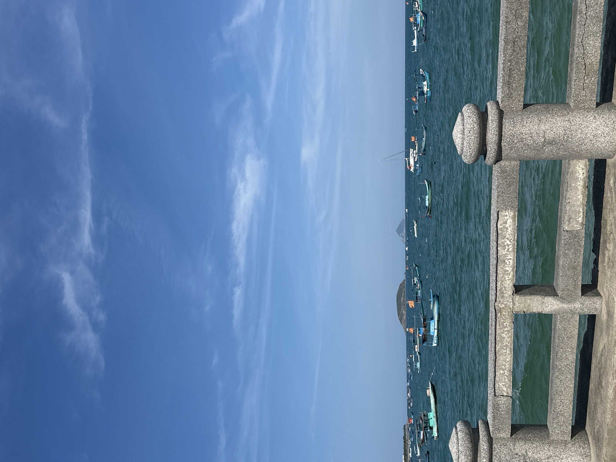 The image depicts a coastal scene in Bac Nha Trang Ward, Vietnam. A concrete pier railing is visible on the right side of the frame. Beyond the railing, numerous boats are anchored in the sea. In the background, land is partially visible. The sky is blue with scattered clouds.
