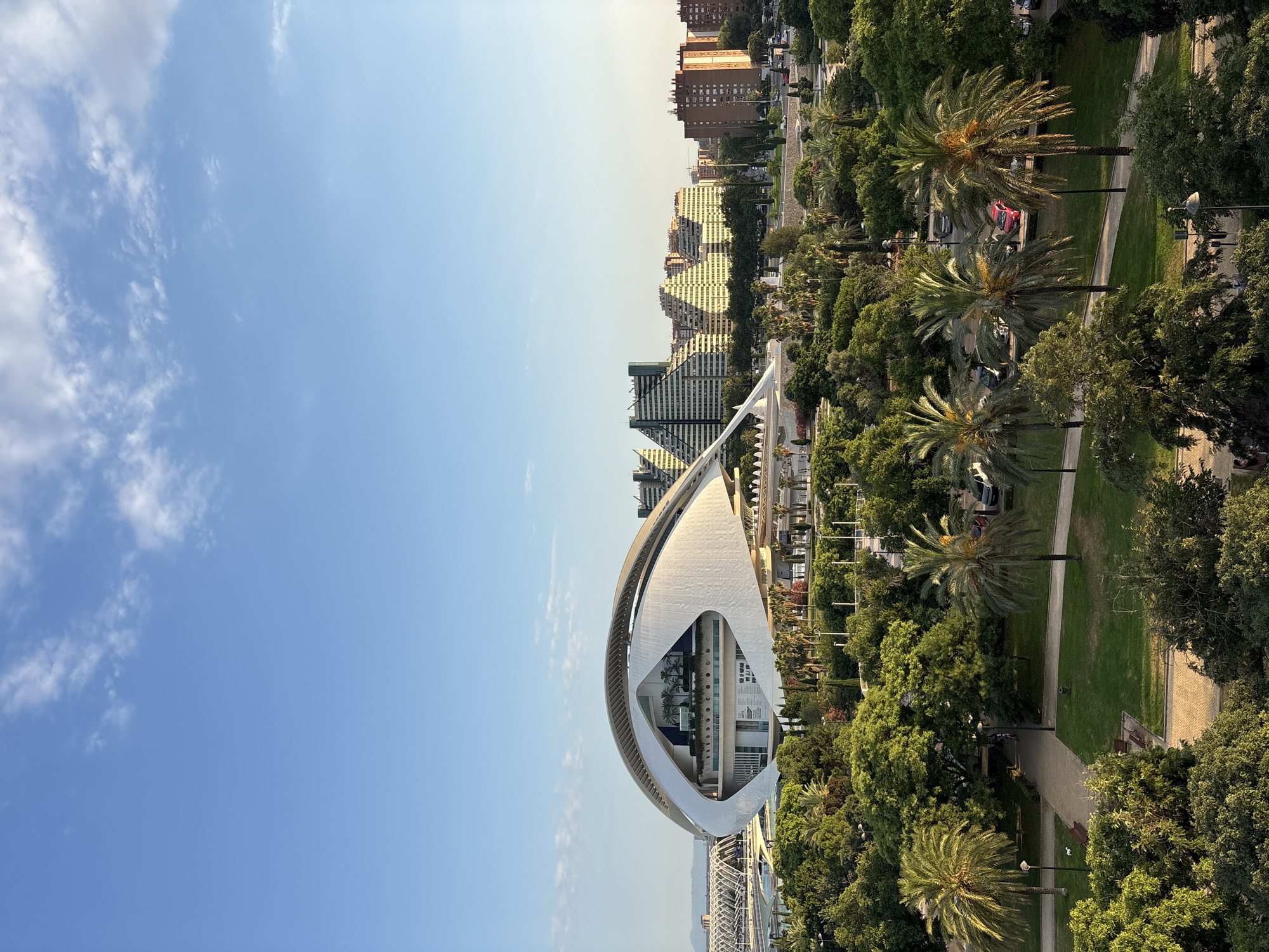 The image captures a panoramic view of the City of Arts and Sciences in València, Spain. In the foreground, lush green trees and palm trees form a vibrant park area with pathways and benches. In the mid-ground, the iconic Hemisfèric, with its distinctive eye-shaped design, stands prominently. Beyond it, several modern buildings with stepped facades, characteristic of the architecture in this area, rise against the clear blue sky. The scene is bathed in the warm light of what appears to be late afternoon or early evening. The sky is a clear blue with scattered white clouds. There are cars parked in the distance, and some distant mountains are visible on the horizon. No people are clearly discernible in the image.