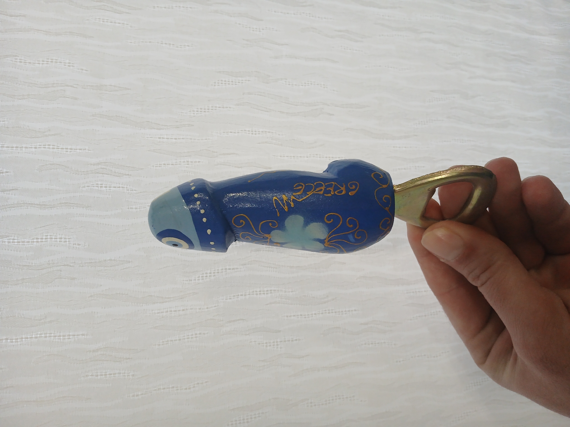 A hand holds a blue phallic-shaped bottle opener, with "GREECE" written on it, against a white, textured background.