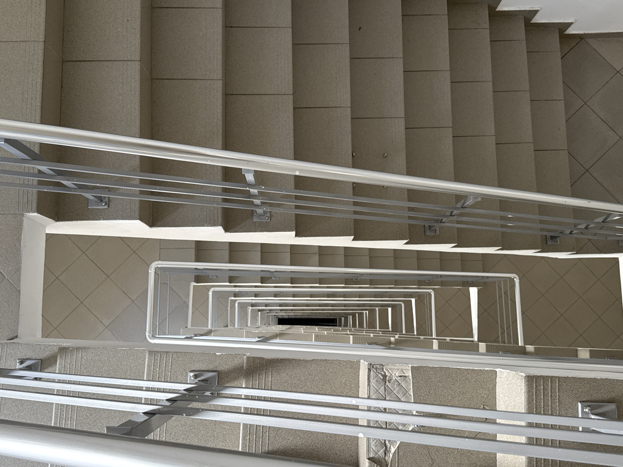 The image shows a top-down view of a staircase in a building, likely located in Sofia, Bulgaria, given the image context. The staircase is constructed of concrete or stone steps, and it winds downwards in a square spiral. Metal handrails with supporting bars run along the inner and outer edges of the staircase. The central opening of the staircase is visible, appearing as a diminishing square tunnel leading into the darkness below. The steps are a light, neutral color, while the handrails are silver. The environment appears clean and well-maintained. The lighting seems natural, possibly from a skylight or windows not visible in the frame. The scene is static, with no people or movement captured.