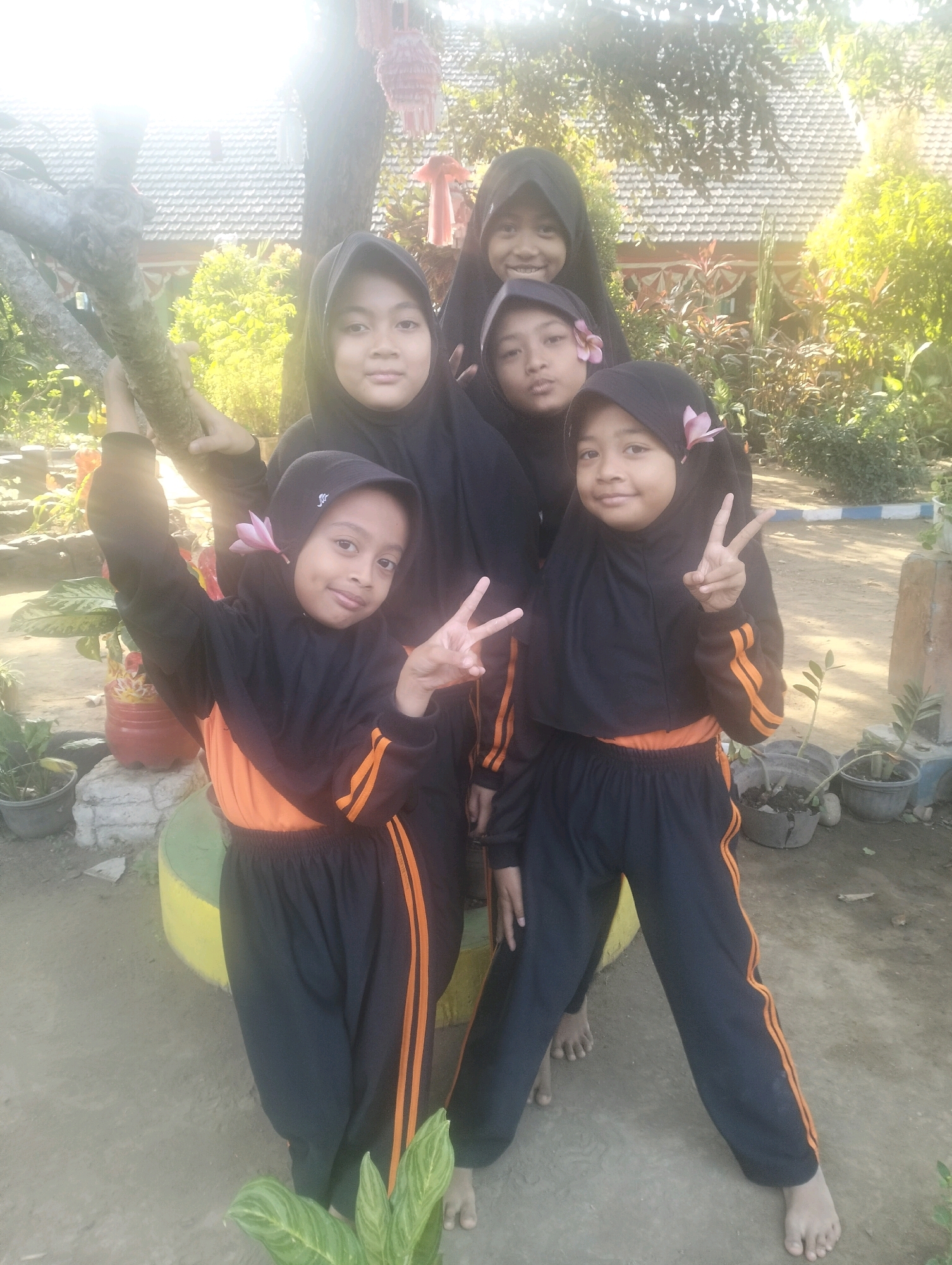 Five young girls wearing black hijabs and orange and black athletic uniforms pose for a photograph outdoors. The girl in the front left is looking at the camera, holding a tree branch with her left hand and making a peace sign with her right hand. She has a pink flower tucked behind her ear. The girl next to her, in the front right, is also making a peace sign with her right hand and looking at the camera. She also has a pink flower behind her ear. The three other girls are in the background, also looking at the camera. They are standing on a dirt path surrounded by greenery, with a building with a tiled roof visible in the background. The image appears to have been taken on a sunny day. The location is identified as Blabakan, Indonesia.