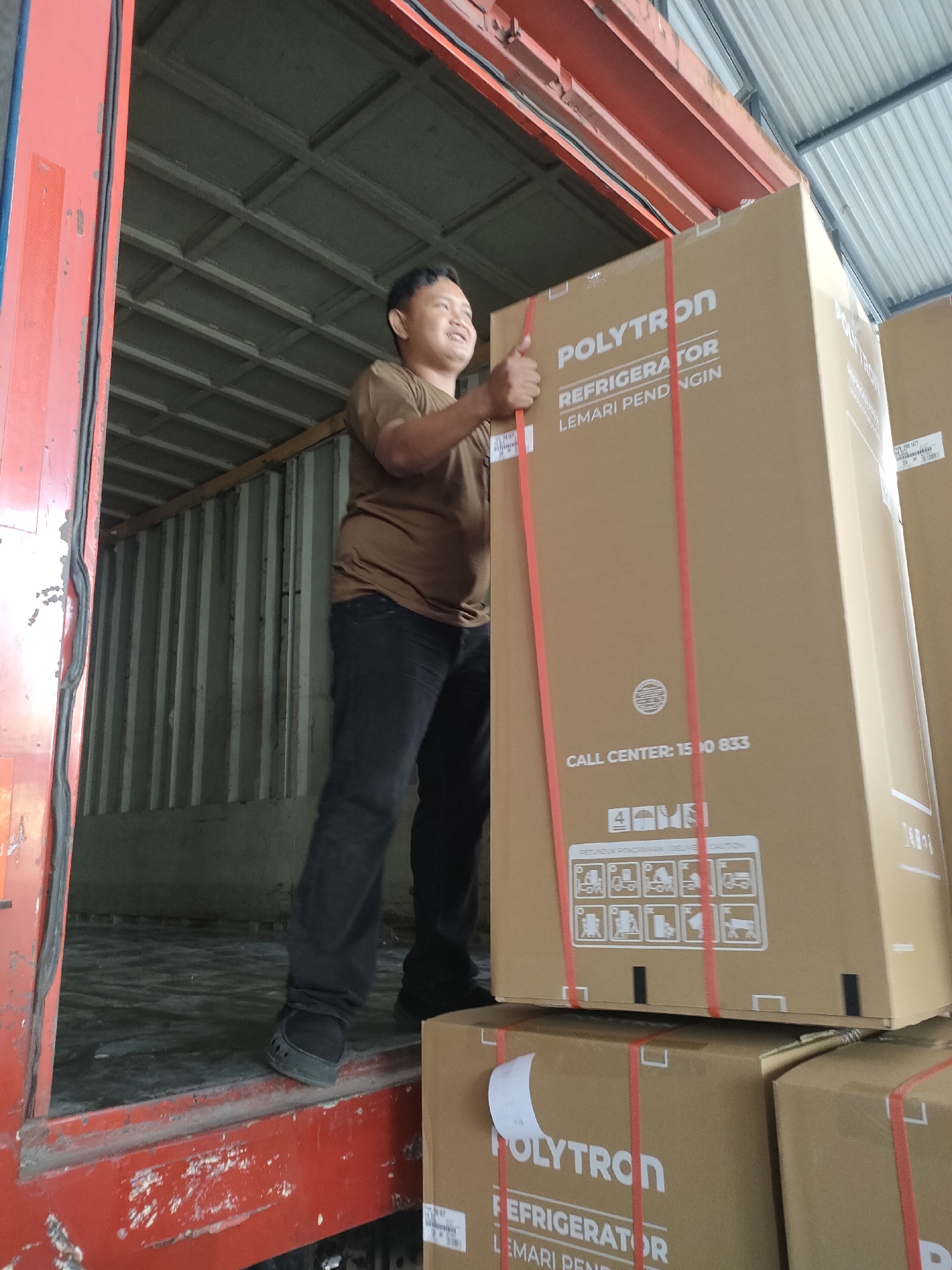 A man in a brown t-shirt and black pants is standing in the open door of a shipping container, holding a large cardboard box with red straps. The box has the word "POLYTRON" and "REFRIGERATOR LEMARI PENDINGIN" printed on it. The man is smiling and giving a thumbs-up. The shipping container appears to be parked in an outdoor industrial area, possibly a warehouse or loading dock, given the corrugated metal walls and roof visible in the background. The overall scene suggests a delivery or loading operation. The provided coordinates place this scene in Sayung, Indonesia, an area known for its industrial and commercial activities, which aligns with the visual cues of the image.
