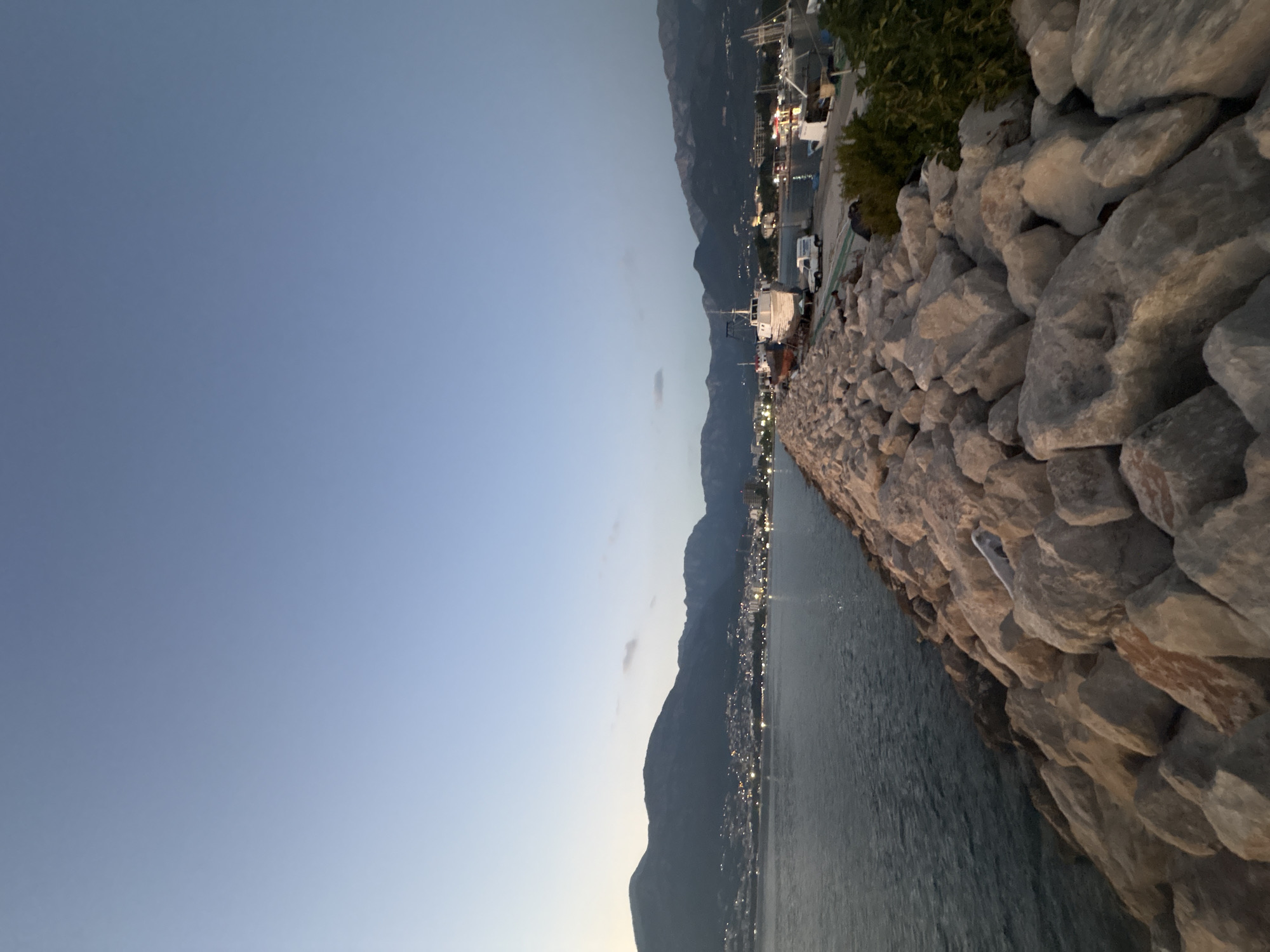 This is a horizontal, eye-level, full shot taken at dusk. The image features a rocky breakwater in the foreground, extending towards the right and into the middle ground, where it meets a body of water. Beyond the water, the city of Bar, Montenegro, is visible along the coastline, with numerous lights twinkling in the twilight. The city is nestled at the base of a mountain range that dominates the background, its peaks silhouetted against the fading light. Several boats are docked in the harbor area of the city. The sky is a clear, pale blue, transitioning to a lighter hue at the horizon, indicating the end of daylight. The water appears calm, with a slight ripple effect.
