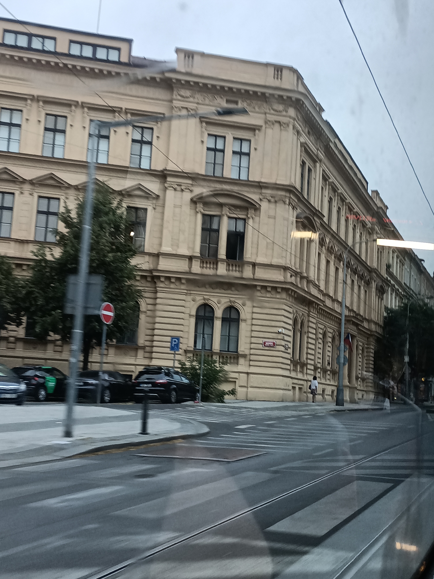 This is a medium shot, taken from inside a vehicle, of a wide, wet street with tram tracks in Prague, Czechia. On the right side of the street is a large, ornate, light-yellow building with multiple floors and many windows. The building has decorative architectural features, including columns and carved details. The street in front of the building is a crosswalk, marked with white stripes, and there are tram tracks running across it. Several cars are parked along the side of the street. One car is dark-colored, and another is bright green. In the background, there is a person walking away from the camera. The sky is overcast and gray.