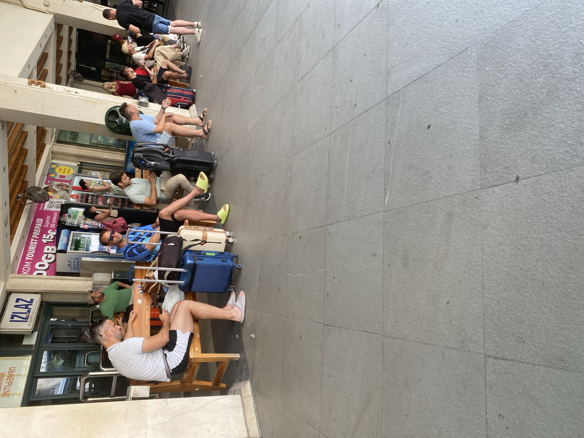 The image shows a group of people sitting on wooden benches in what appears to be a waiting area, likely at a bus or train station, given the luggage present. The people are diverse in age and gender, and they are all dressed casually. Some are looking at their phones, while others are talking or resting. The scene is set outdoors, with a tiled floor and a building with large windows in the background. There are several pieces of luggage, including suitcases and backpacks, placed near the benches.
Visible text in the image includes "IZLAZ" which means "exit" in Serbian, and "KOM TOURIST PREPAID DOGB 15€ Superior 5G network" suggesting a tourist prepaid mobile service. The overall mood appears relaxed, with people waiting patiently. The lighting suggests it is daytime, and the weather seems pleasant. The location is identified as Budva, Montenegro, which is consistent with the presence of Serbian language text and a tourist-oriented prepaid service. The scene conveys a sense of travel and waiting.