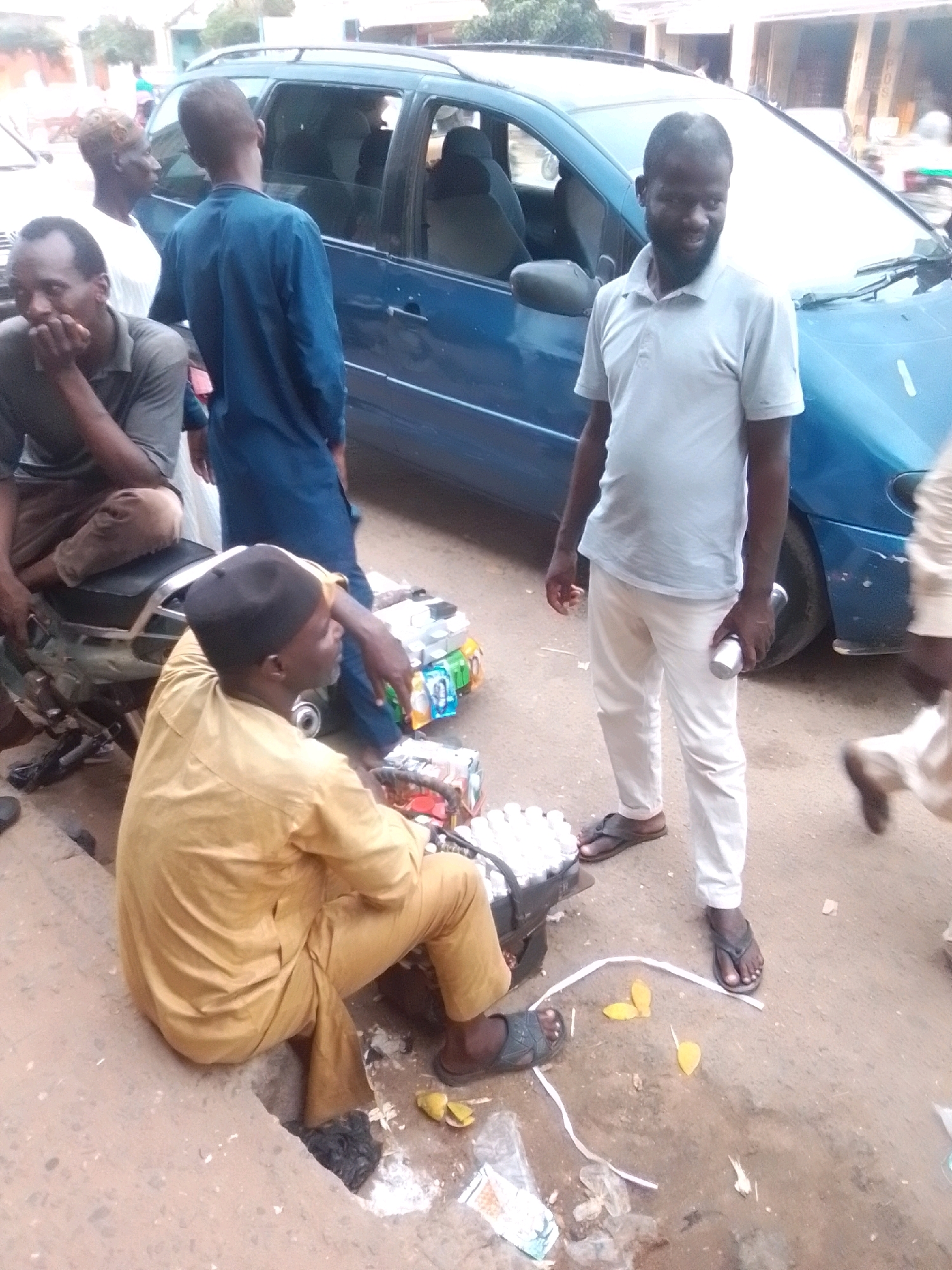 In Arawa, Nigeria, a group of men are gathered near a blue minivan. One man, wearing a light blue polo shirt and white pants, stands with a silver cylinder in his right hand, looking towards the camera with a slight smile. Another man, in a mustard-yellow traditional garment and black cap, sits on the ground, looking to his right, with a pile of packaged goods and a motorcycle behind him. A third man, in a dark blue garment, stands near the minivan's open door, his back to the camera. A fourth man, wearing a grey shirt and brown pants, sits on the motorcycle, looking down with his hand to his chin. The scene appears to be an outdoor market or roadside stall, with various packaged items scattered on the ground. The environment is dusty and appears to be a typical street scene in the city.