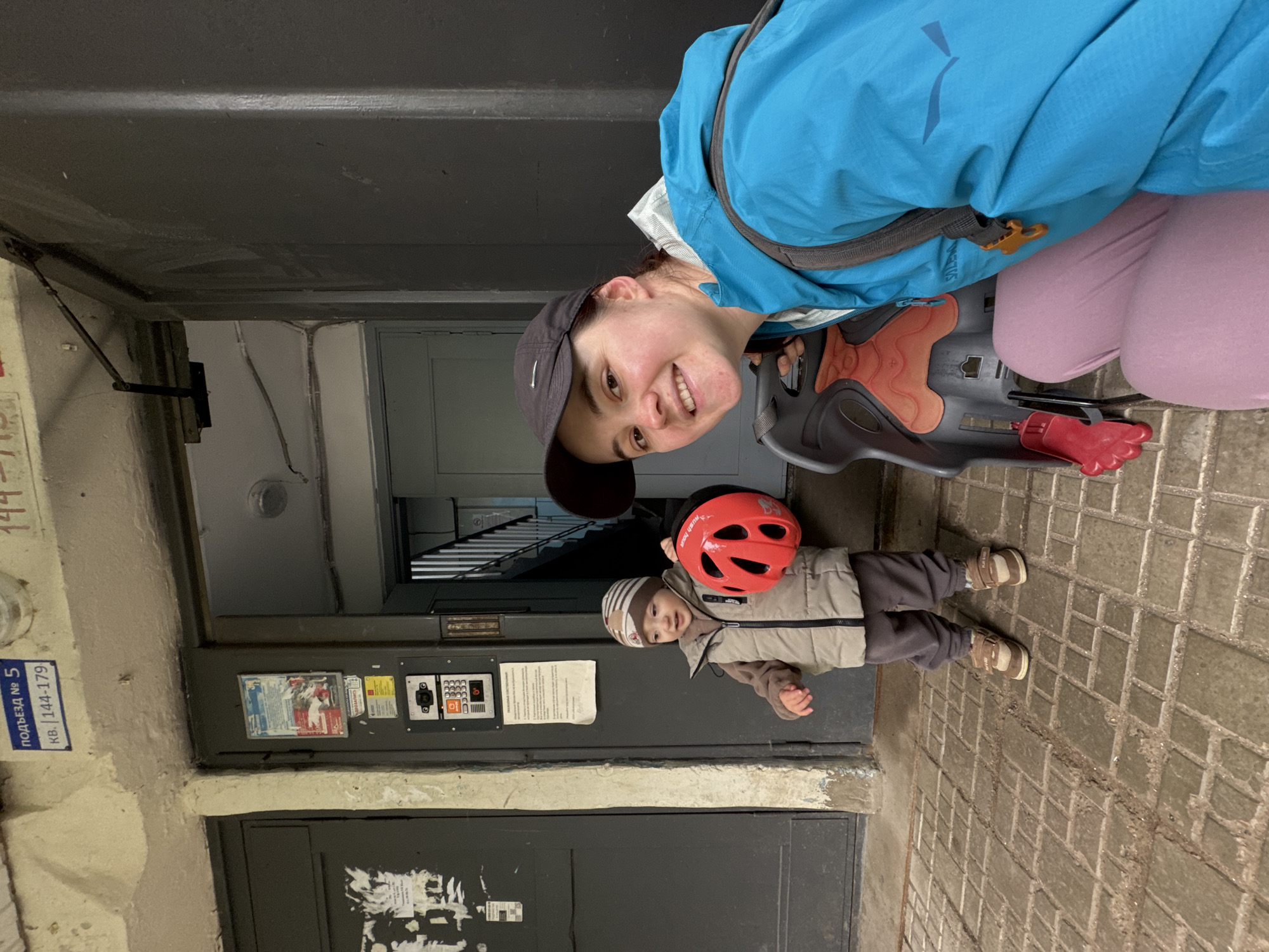 A woman and a young child stand outside an apartment building entrance in Izhevsk, Russia. The woman, wearing a blue jacket and a black baseball cap, smiles at the camera while holding a child's bicycle helmet. A child's bicycle seat is visible beside her. The child, dressed in a puffer vest and a striped hat, stands to the left of the woman, holding the red helmet. The entrance of the building features a gray door, an intercom system, and a sign indicating "ПОДЪЕЗД № 5" (Entrance No. 5). The ground is paved with interlocking bricks.