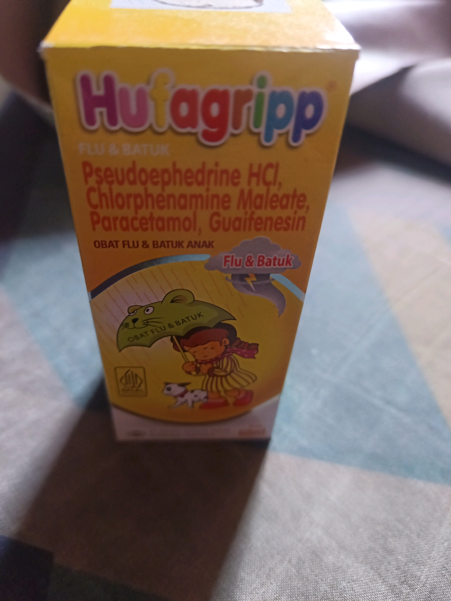 A yellow cardboard box for a children's cough and cold medicine called "Hutagripp" is positioned on a patterned surface, likely a bed or tablecloth, in what appears to be an indoor setting. The box displays the brand name in colorful lettering, followed by "FLU & BATUK" (Flu & Cough in Indonesian). Below this, the active ingredients are listed: "Pseudoephedrine HCl, Chlorphenamine Maleate, Paracetamol, Guaifenesin." Further down, it specifies "OBAT FLU & BATUK ANAK" (Children's Flu & Cough Medicine). The box features illustrations of a cartoon rat holding an umbrella that reads "OBAT FLU & BATUK," a child in a raincoat and boots, and a small dog. A halal certification logo is visible on the lower left side of the box. The lighting suggests a natural light source, possibly from a window, casting a shadow to the lower right of the box. The background is out of focus, showing a textured surface with stripes of blue, white, and beige. There is no visible indication of a specific location within South Jakarta, Indonesia.