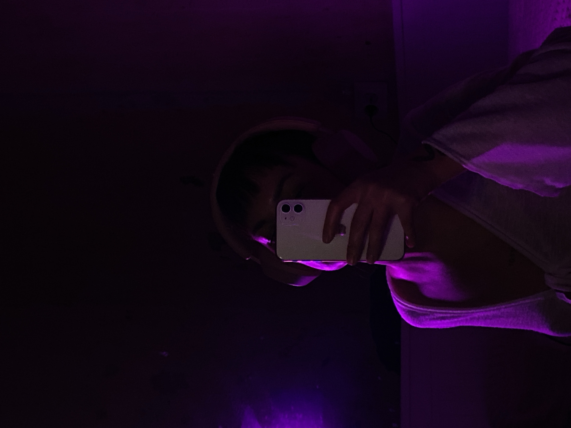 A person with dark hair is taking a mirror selfie in a dimly lit room with purple lighting. The person is holding a white smartphone, obscuring part of their face. They are wearing a light-colored hooded sweatshirt or robe. The environment appears to be a room with dark walls and minimal discernible details aside from an electrical outlet on the wall near the top right. The presence of a mirror suggests the location is likely an interior space like a bathroom or bedroom. It's impossible to determine if this scene occurred in Votkinsk, Russia, as there is no contextual information in the image to suggest the location.