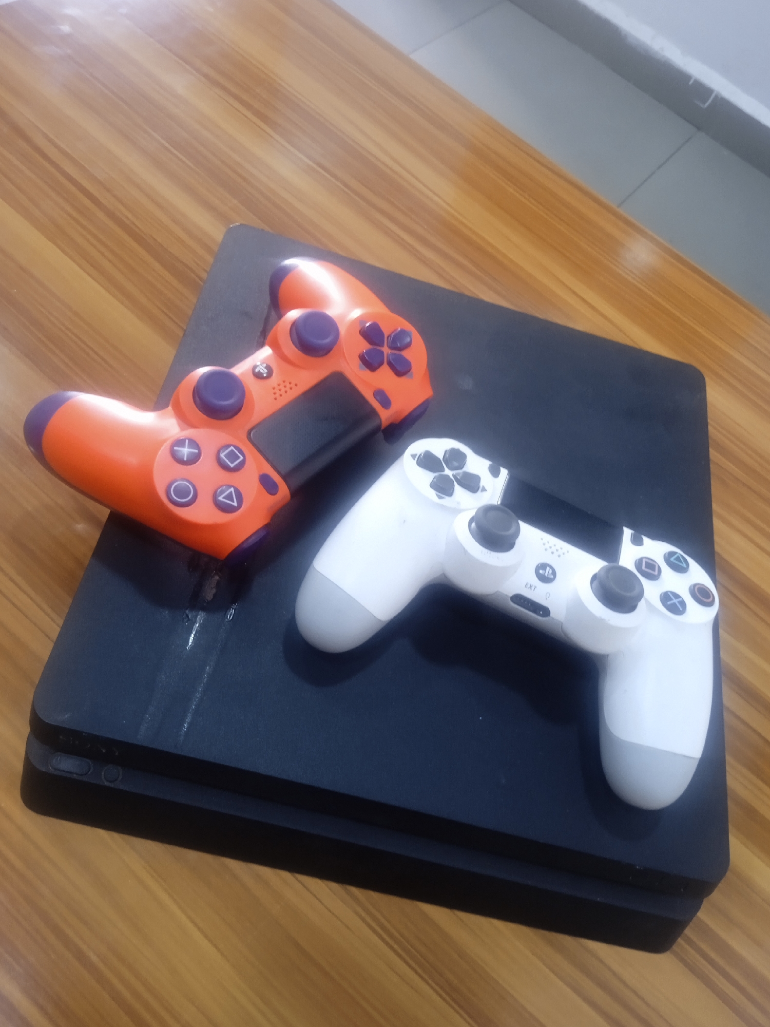 The image captures a black PlayStation 4 console lying on a light brown wooden table. Two PlayStation 4 DualShock controllers are resting on top of the console. The setting appears to be indoors, likely in a room in Jos, Nigeria.
The controller on the left is a vibrant orange with dark purple accents on its thumbsticks, directional pad, and face buttons (X, O, Square, Triangle). The controller on the right is white with light grey thumbsticks and grips, also featuring the standard PlayStation button symbols. Visible text on the white controller includes "EXT" and the PlayStation logo. Both controllers show signs of use but appear to be in functional condition.
The PlayStation 4 console has a matte black finish, with some visible dust or minor smudges on its surface. The wooden table exhibits a distinct grain pattern. In the upper right background, a light-colored wall or tiled floor is visible. The overall lighting is even, suggesting either natural daylight or consistent artificial illumination, without indicating a specific time of day. No people are present, and no activity is explicitly taking place; the items are simply arranged on the table.