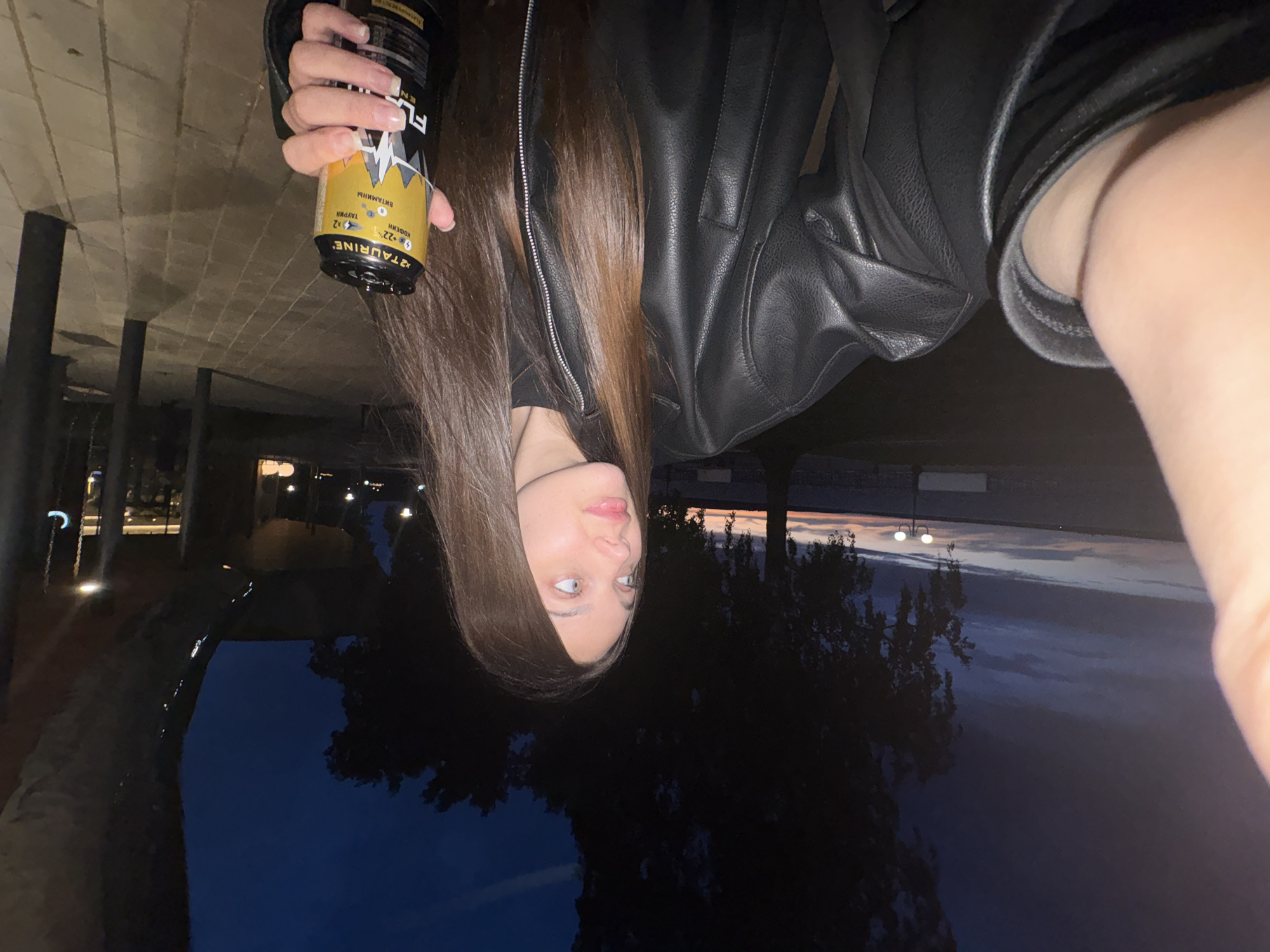 The image shows a selfie taken by a female with long brown hair wearing a black leather jacket and holding a can of energy drink. She is outdoors, likely at night, under a structure that might be an overpass or similar architectural feature. The background shows dark silhouettes of trees and what seems to be a twilight sky. The location is suggested to be Votkinsk, Russia, but there's no visual confirmation. The can is partially obscured, but it appears to be a yellow and black energy drink with some text in a language other than English. The lighting is dim, contributing to the overall low-light condition of the image.