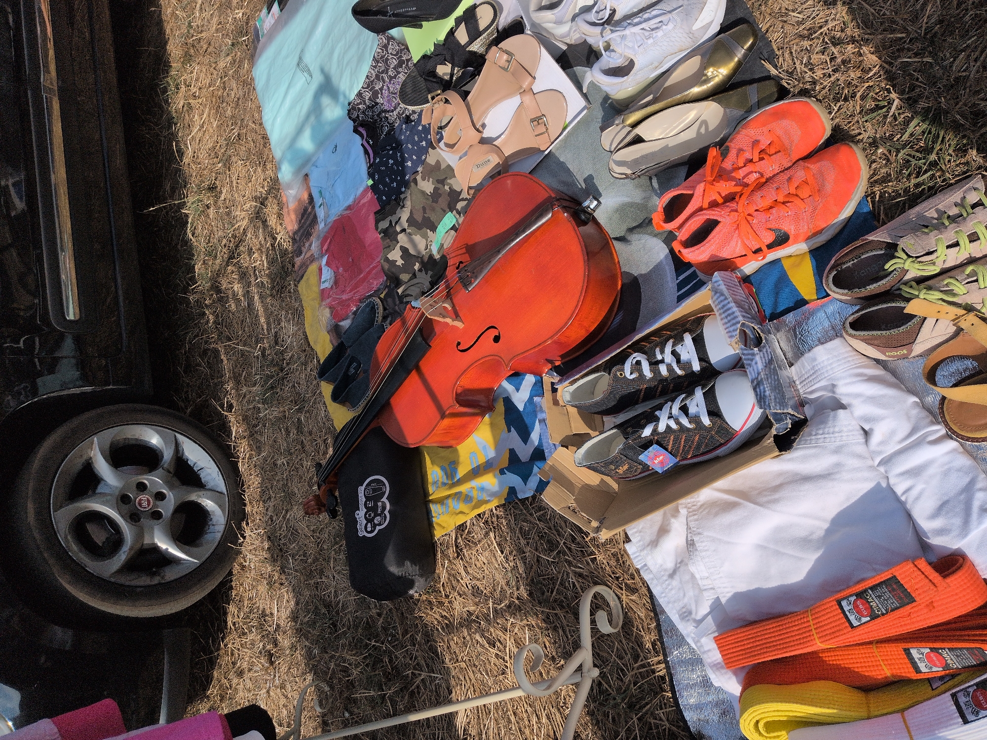 The image shows a collection of items laid out on the ground, likely at a car boot sale or flea market, with a car visible in the background. The scene is set in the city of South Staffordshire, United Kingdom.
The items include:
- A bright orange cello or large violin.
- A black cello case with a white logo.
- Various clothing items, including t-shirts, patterned fabric, and a camouflage fabric.
- Several pairs of shoes, including sandals, sneakers, and formal shoes.
- Three martial arts belts, one yellow and two orange, with visible labels.
- A white garment, possibly a gi or a shirt.
- A white metal stand with a decorative scroll design.
- The rear wheel of a black car.
- The ground is covered in dry grass and straw.
The lighting suggests it is daytime, with shadows cast by the objects. The overall impression is of items for sale or display in an outdoor market setting.