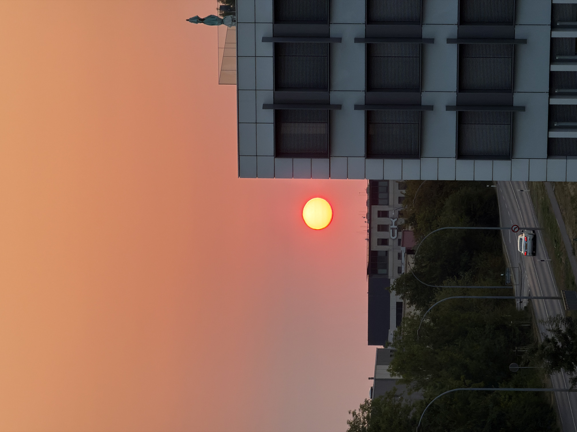 The image depicts a sunset over a city, likely Leudelange, Luxembourg, with a modern office building in the foreground. The sky is a vibrant orange and pink gradient, with the setting sun appearing as a bright, reddish-yellow orb. The office building is a multi-story structure with a facade of light-colored panels and dark-framed windows. A statue, possibly of the Virgin Mary, stands on the rooftop of the building. In the background, other buildings and lush green trees are visible. A road with a white car driving on it runs in front of the trees. Streetlights line the road, and a traffic sign is visible. The overall scene suggests an urban environment at dusk.