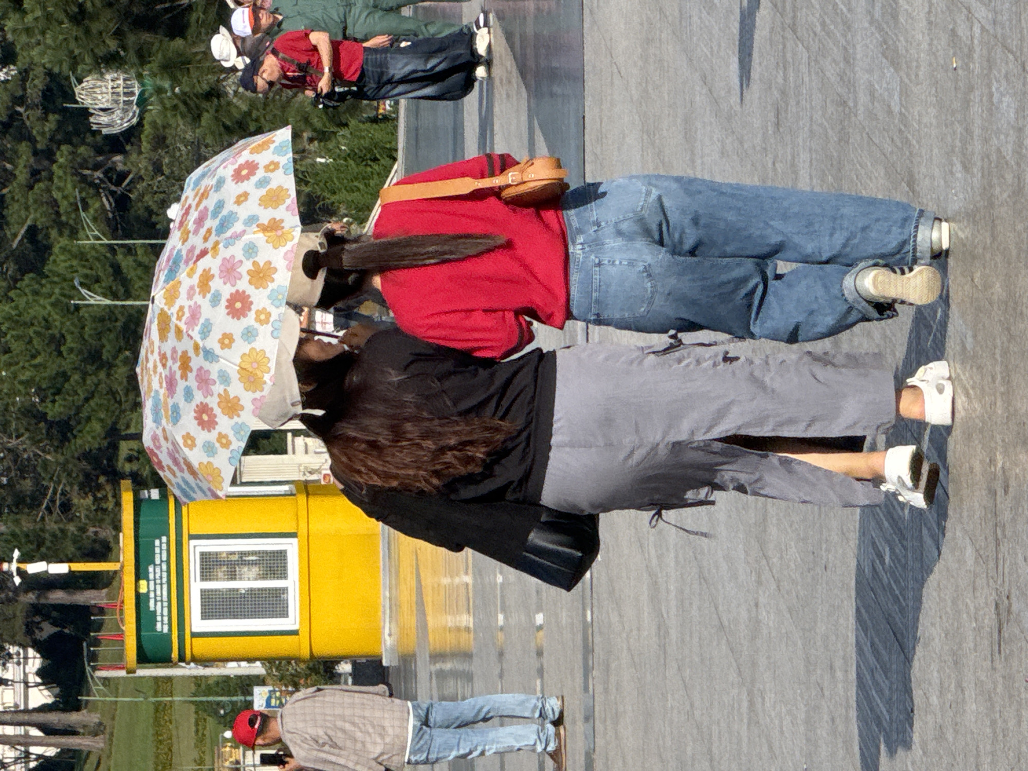 Two young women walk side-by-side under a colorful floral umbrella in Da Lat, Vietnam. The woman on the left wears a black long-sleeved shirt, a grey maxi skirt, and white sneakers. She carries a black handbag. The woman on the right wears a red long-sleeved shirt, blue jeans, and white sneakers. She has a tan crossbody bag. In the background, a yellow ticket booth is visible, and several other people are scattered around the plaza. The ground is paved with grey tiles, and trees line the background. The scene appears to be a public space, possibly a park or tourist attraction.