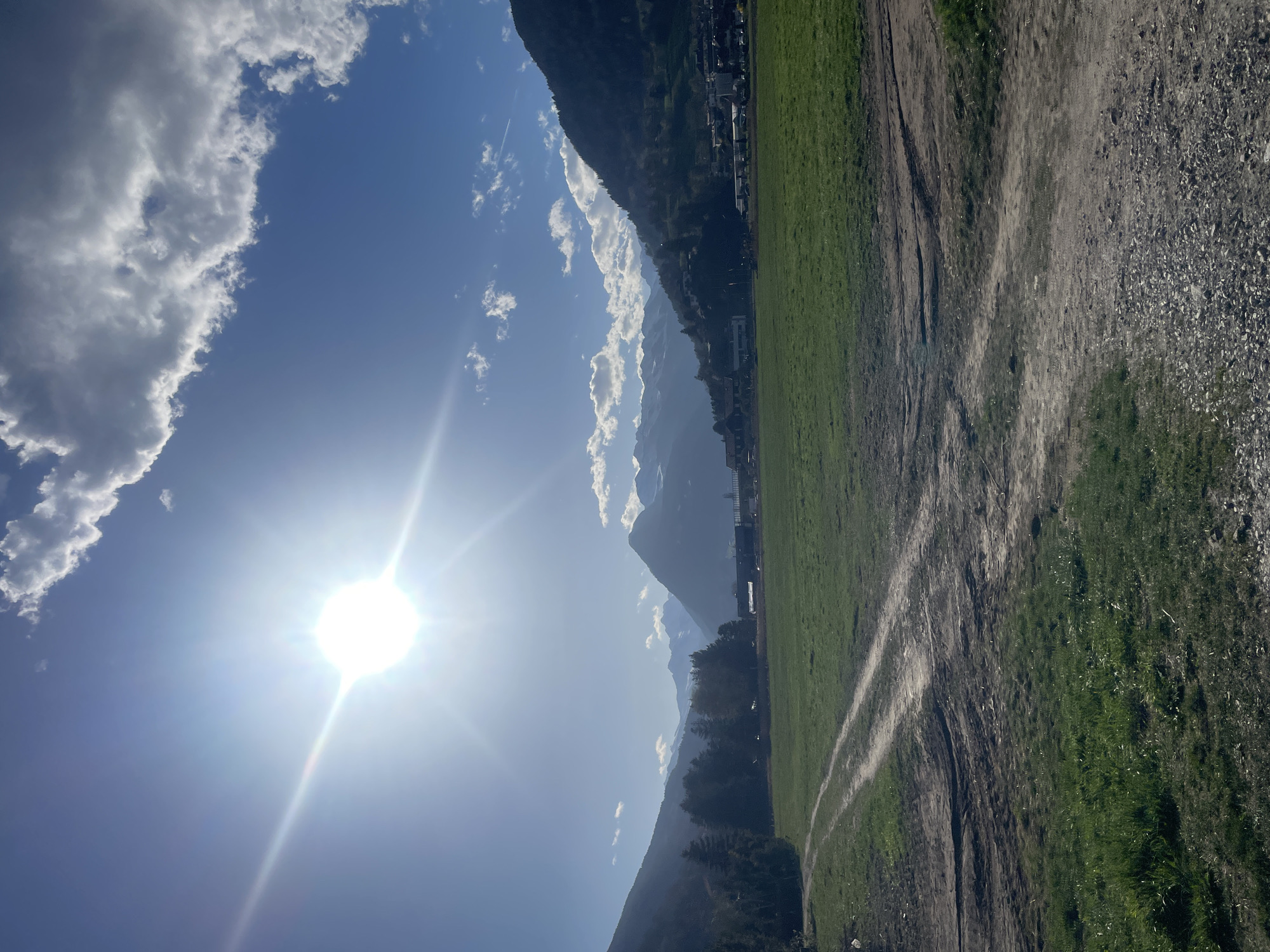 The image presents a landscape scene in Sterzing-Vipiteno, Italy, under a bright sun. The sky is blue with scattered white clouds. The sun's glare dominates the left side of the image. Mountains are visible in the background, partially obscured by clouds. In the foreground, there's a field of green grass. Adjacent to the grass, a dirt path or road is visible, indicating some level of traffic. The image appears to be taken during daylight hours.