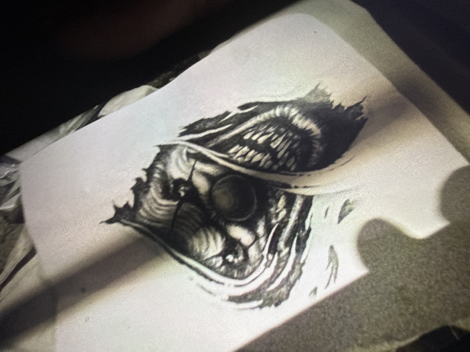 The image displays a close-up, angled view of a sketch on white paper. The sketch depicts a stylized, possibly demonic or monstrous face with sharp teeth and a large, dark eye. The rendering is in black and white, utilizing shading to create depth and texture. Shadows obscure parts of the paper and the surrounding environment, suggesting an indoor setting with limited, directional lighting. The exact location in Sofia, Bulgaria, cannot be determined from the visual information provided.