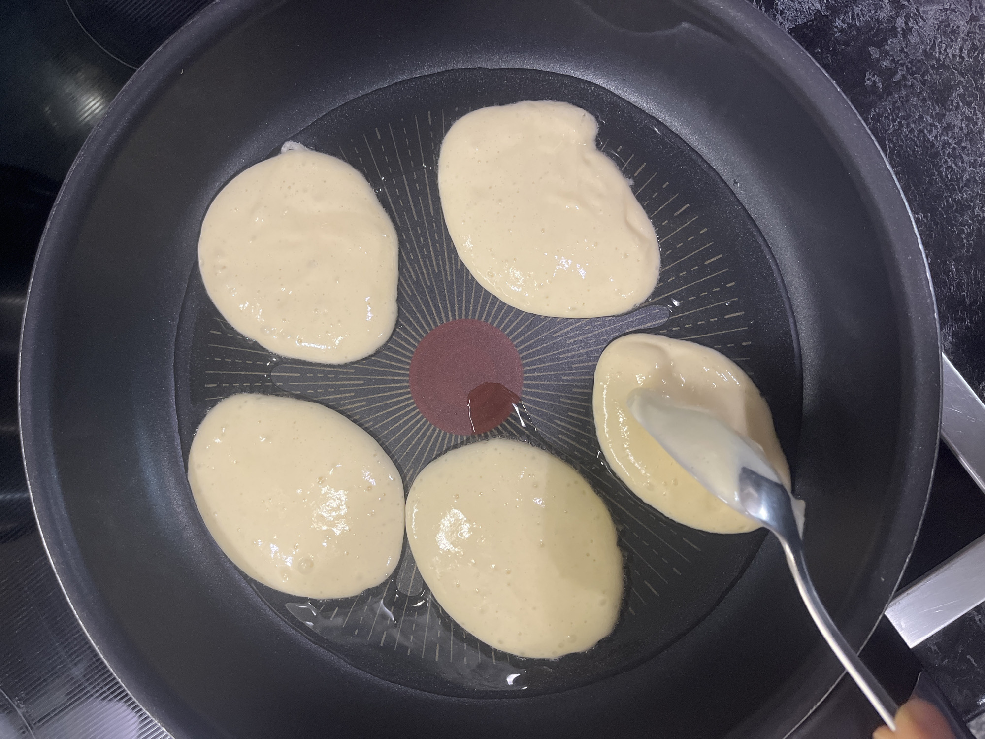 A top-down shot shows five small pancakes cooking in a black non-stick frying pan. A stainless steel spoon is held above one pancake, apparently adding more batter. The pan sits on a stovetop. The location is not discernible from the image.