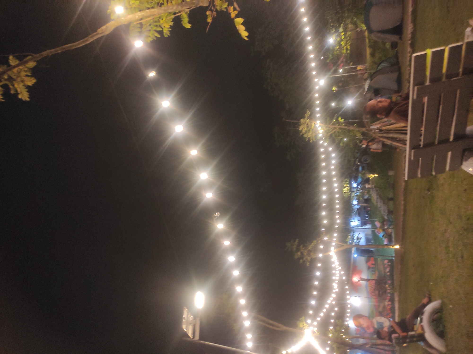 At night in Babakan Madang, Indonesia, a line of glowing string lights stretches into the distance. A tree branch with leaves is visible on the left. On the right, there's grass, and a person is sitting. Beyond them, there's a tent. Further back, a colorful wall painting can be seen.