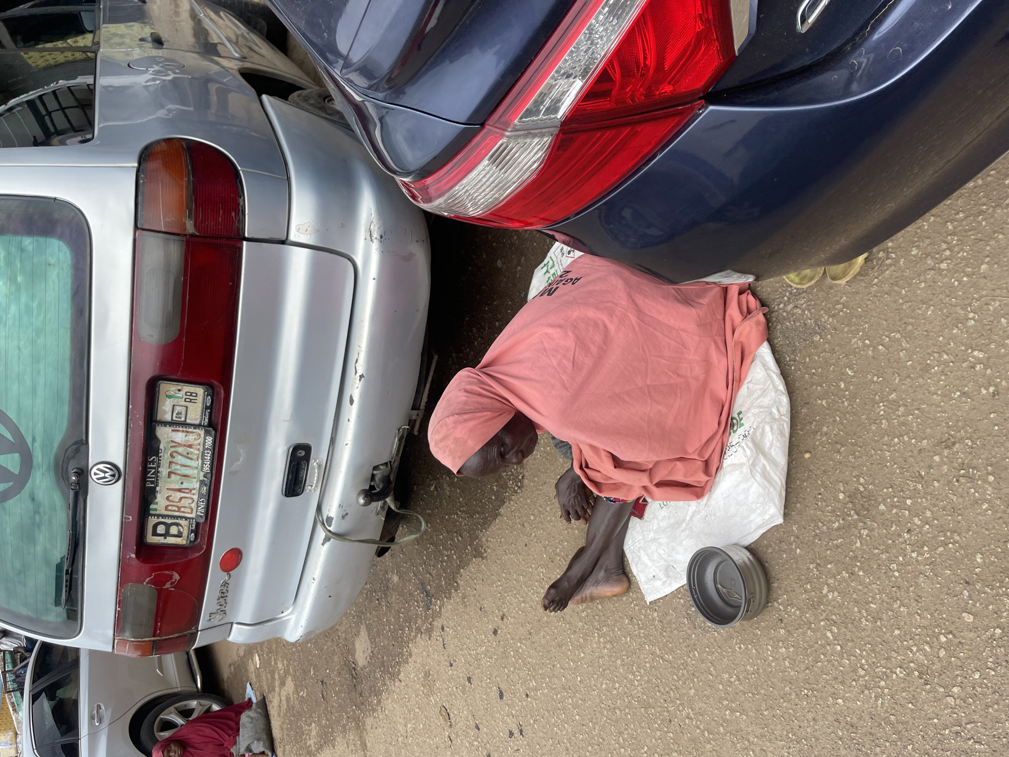 A street scene in Jos, Nigeria, depicts an elderly woman in a pink headscarf and wrap seated on a white cloth on the ground, with a metal bowl in front of her. She is positioned next to a dark blue car and a silver minivan, with the minivan's license plate visible as "BSA-772XJ". The ground is paved with asphalt.