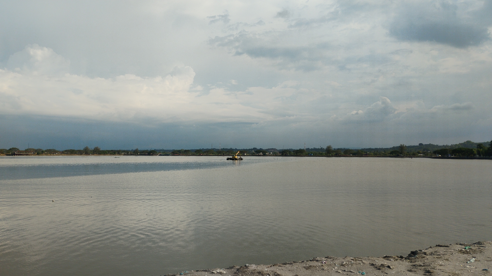 A wide, gray body of water dominates the foreground and midground of the image. The water has a slightly rippled texture, indicating gentle movement. In the distance, along the horizon, is a low-lying landmass covered with trees and some structures, suggesting a coastal area or a town. The sky above is overcast with a mix of light and dark clouds, with the darker clouds creating a moody, diffused light. In the middle of the water, a yellow excavator is operating from a pontoon, its arm extended as it works. The overall impression is a somewhat industrial or construction scene in a natural, watery environment. This scene is consistent with the landscape near the mangrove forest areas of Lhokseumawe, Indonesia, where such waterways and construction activities are common.